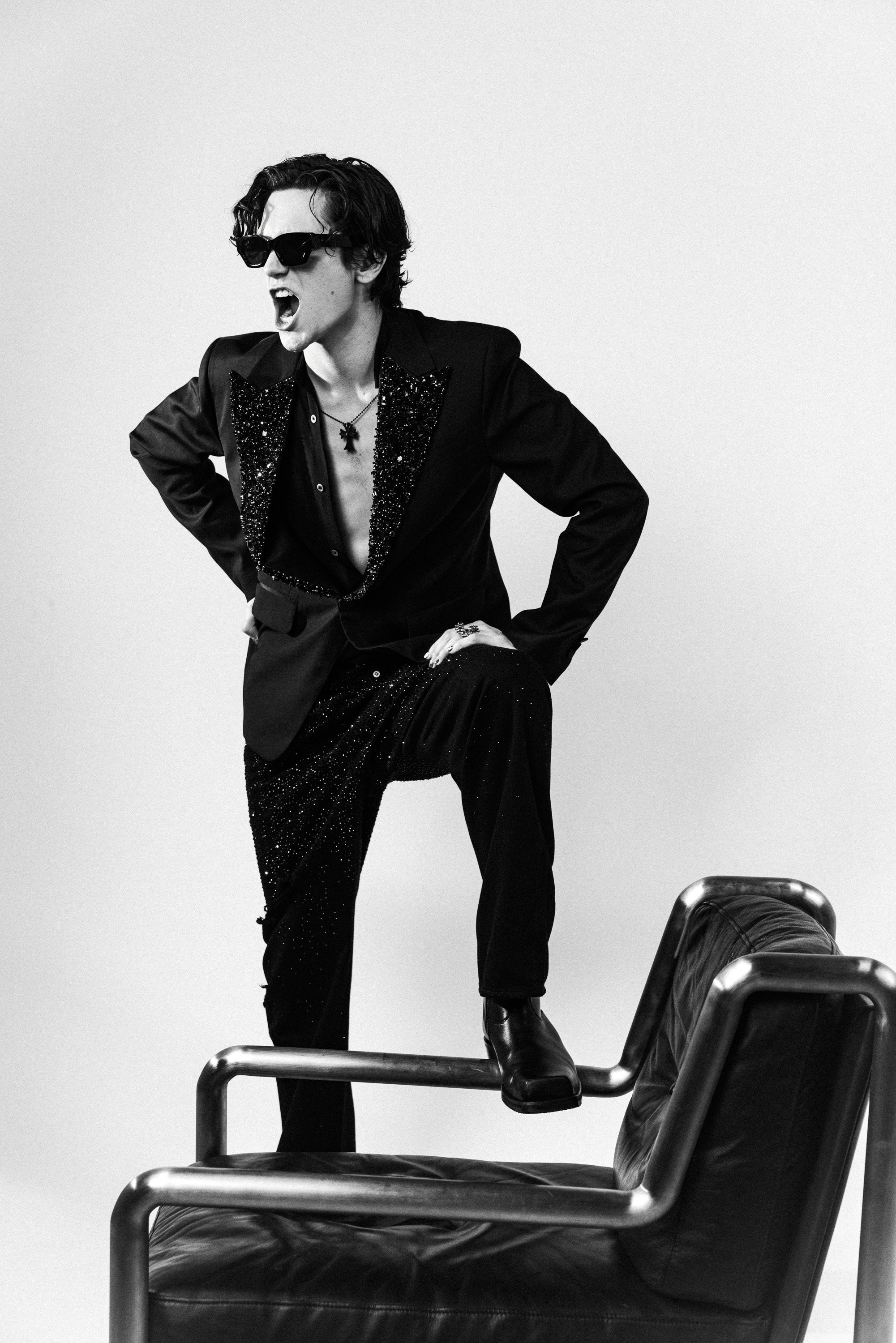 Black and white photo of a man with dark hair, wearing sunglasses and a suit with a sparkly lapel, standing with hands on hips and yelling, while balancing one foot on a leather armchair.