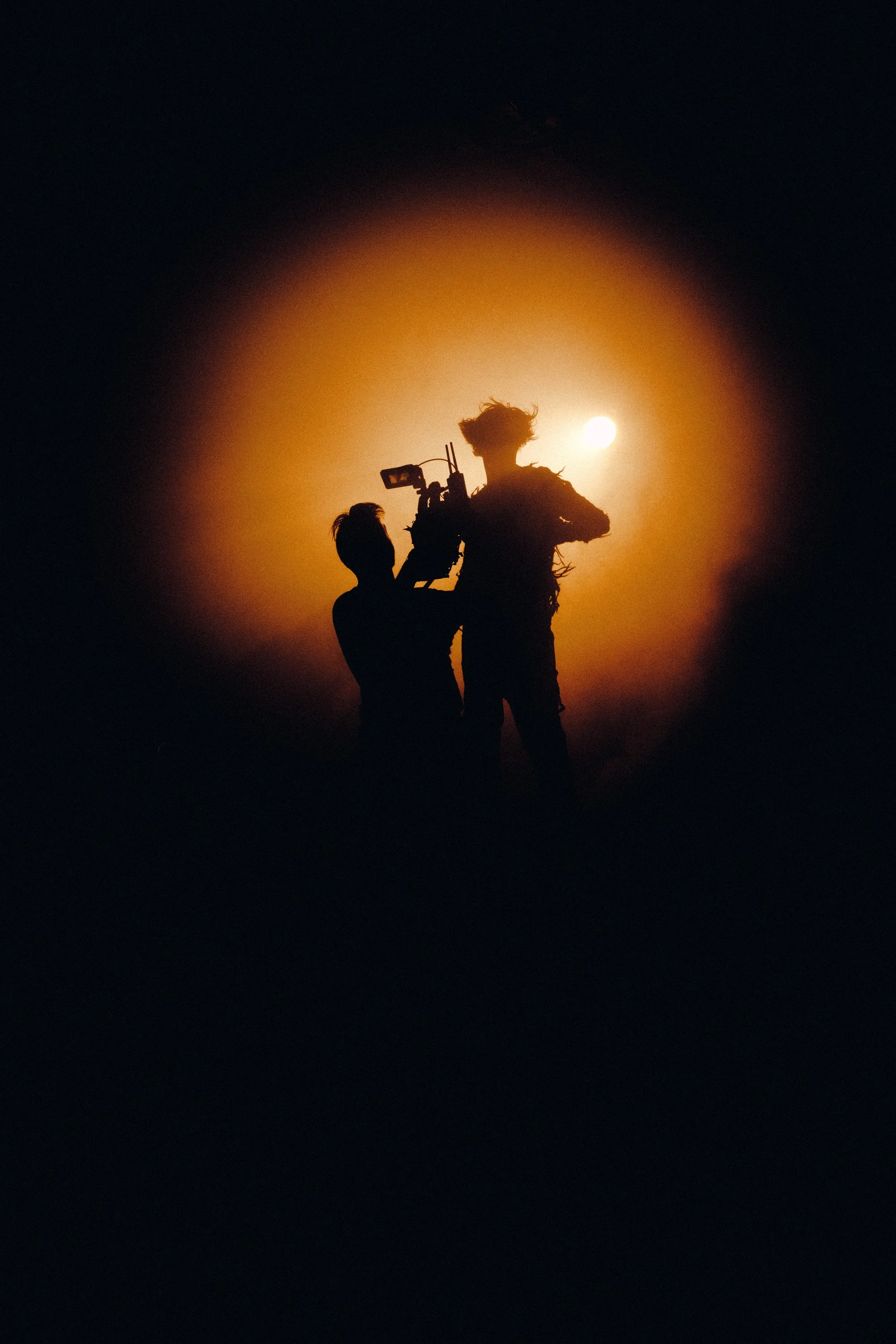 Silhouettes of two people, one holding a camera, against a bright sun in a dark sky at sunset or sunrise.