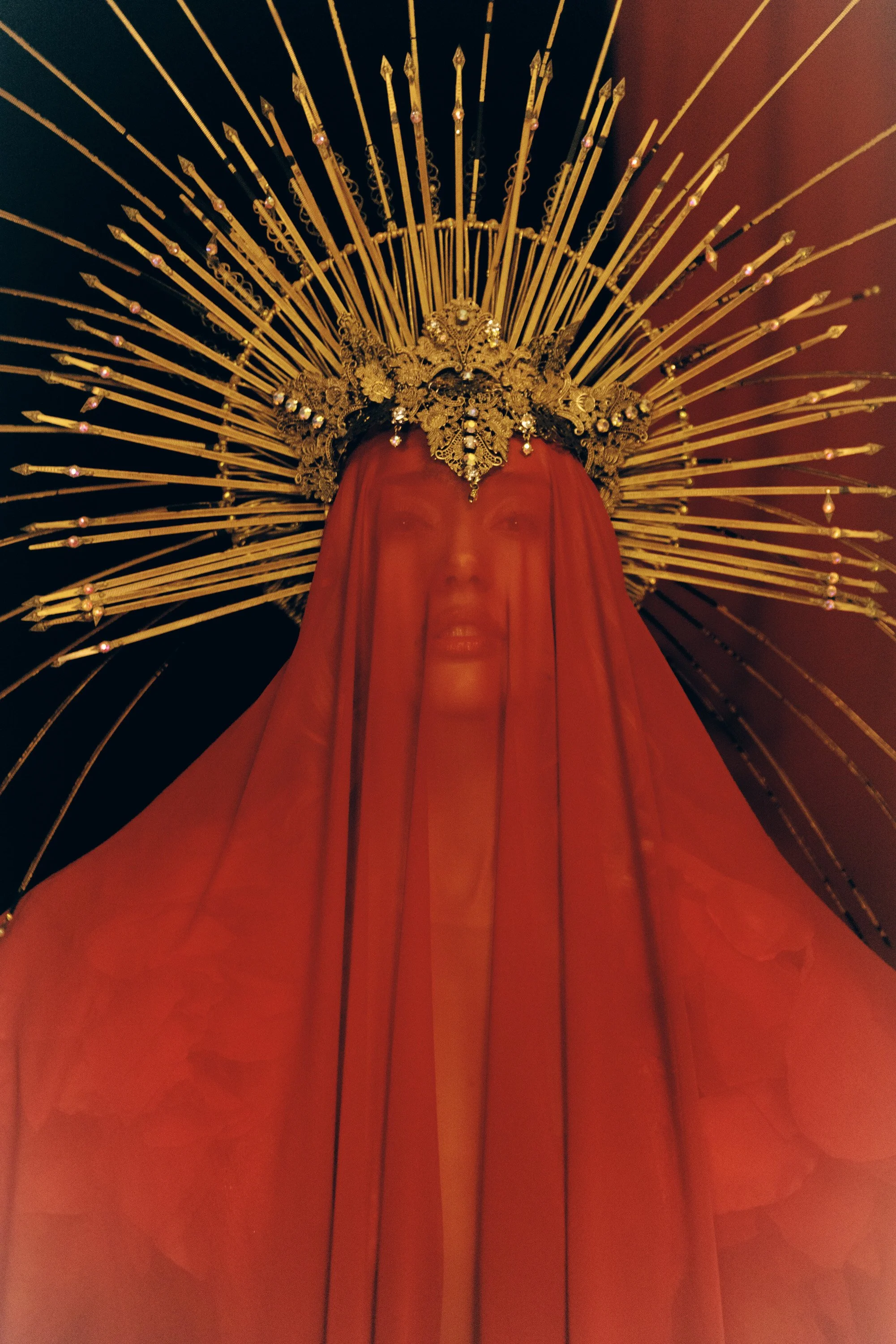 A religious statue of the Virgin Mary with a gold crown and a red veil, behind which radiate multiple golden spikes.