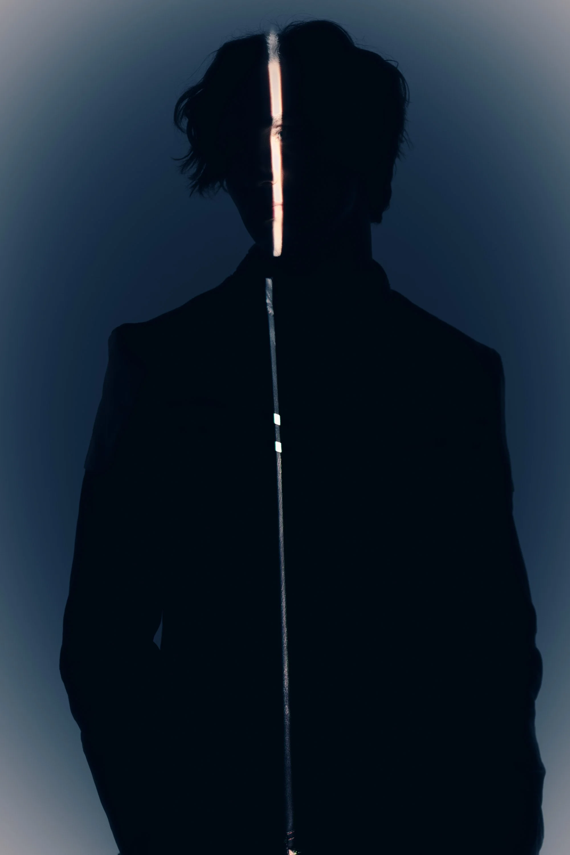 A person with dark, wavy hair silhouetted against a dark background, with a vertical strip of bright light illuminating their face along the center.