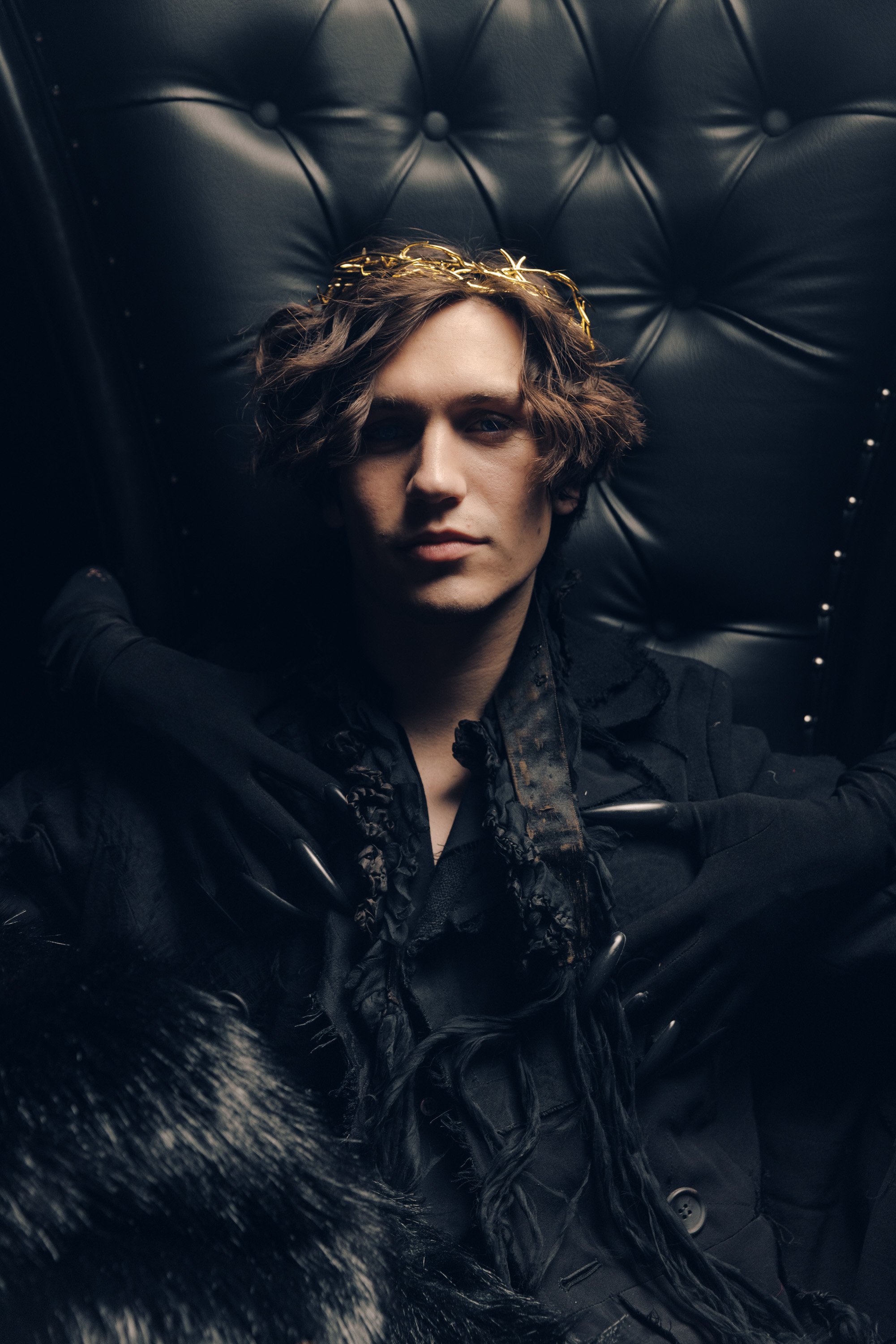A young man with medium-length wavy hair, wearing a black outfit with ruffled details, sitting against a tufted black leather background, wearing a gold crown or headpiece with a subtle confident expression.
