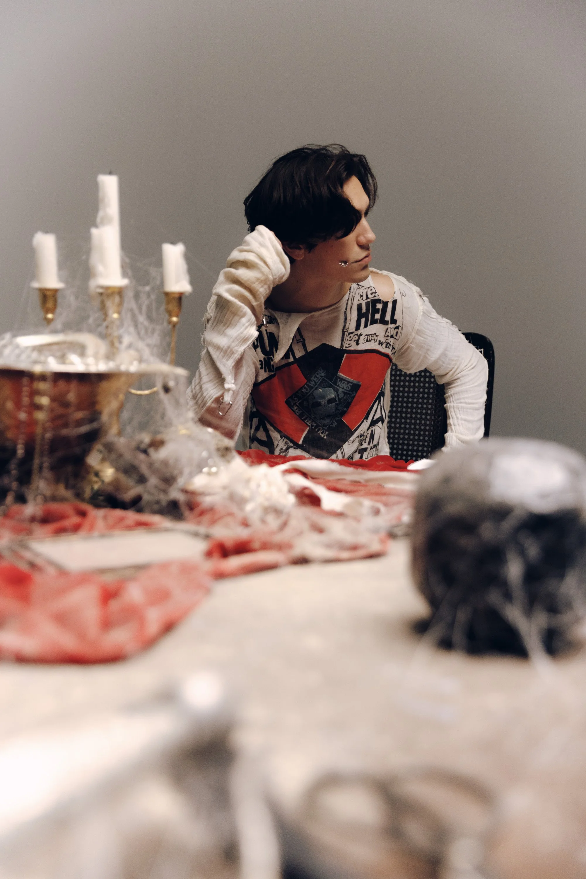 A young man with dark hair, wearing a graphic long-sleeve shirt, seated at a table decorated with Halloween-themed items including a candelabra with white candles, fake cobwebs, and Halloween decorations, in an indoor setting with plain walls.