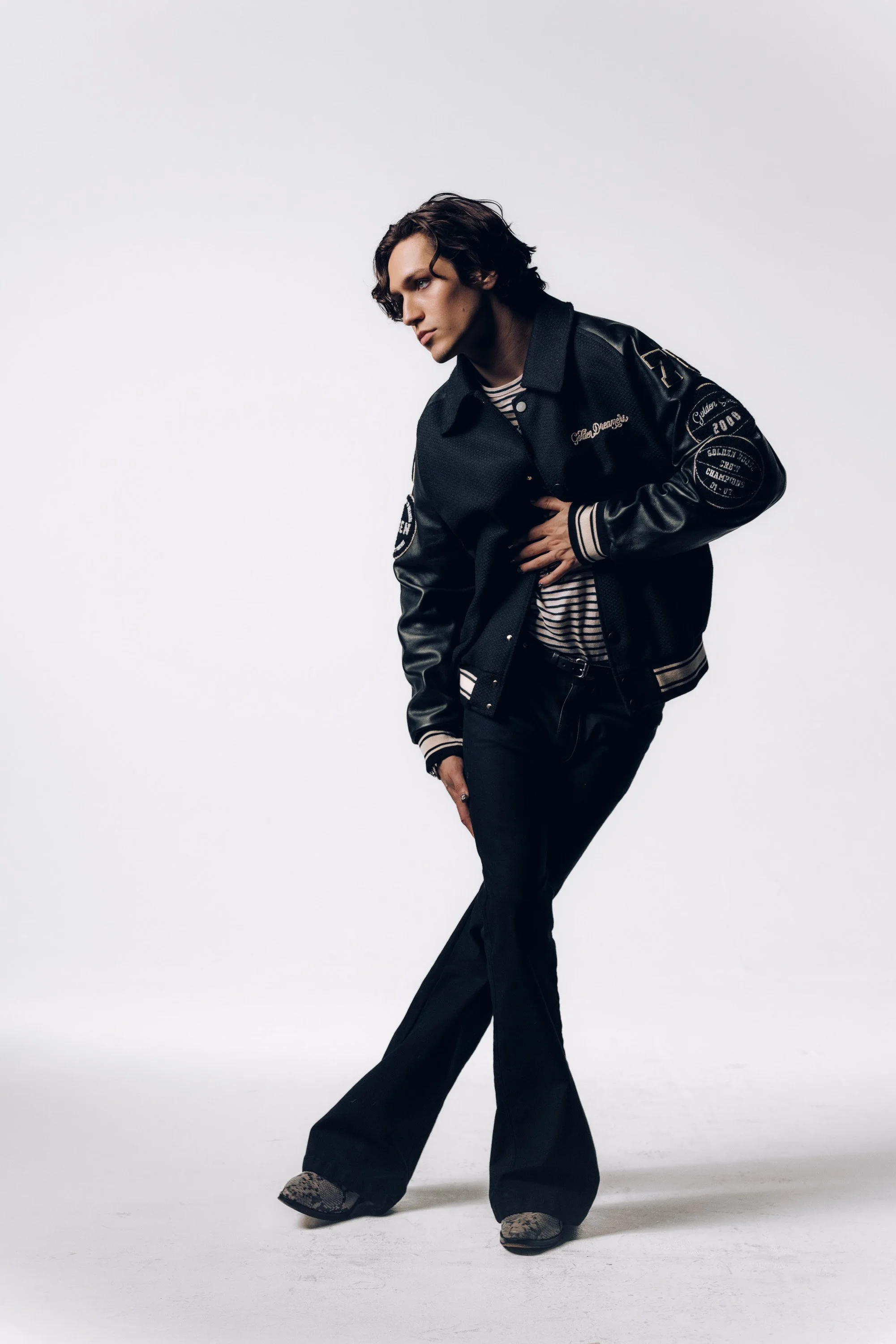 A young man with wavy dark hair and light skin is standing against a plain white background. He is wearing a black varsity jacket with patches and embroidery, a striped shirt underneath, black wide-legged pants, and snake print shoes. He is leaning f