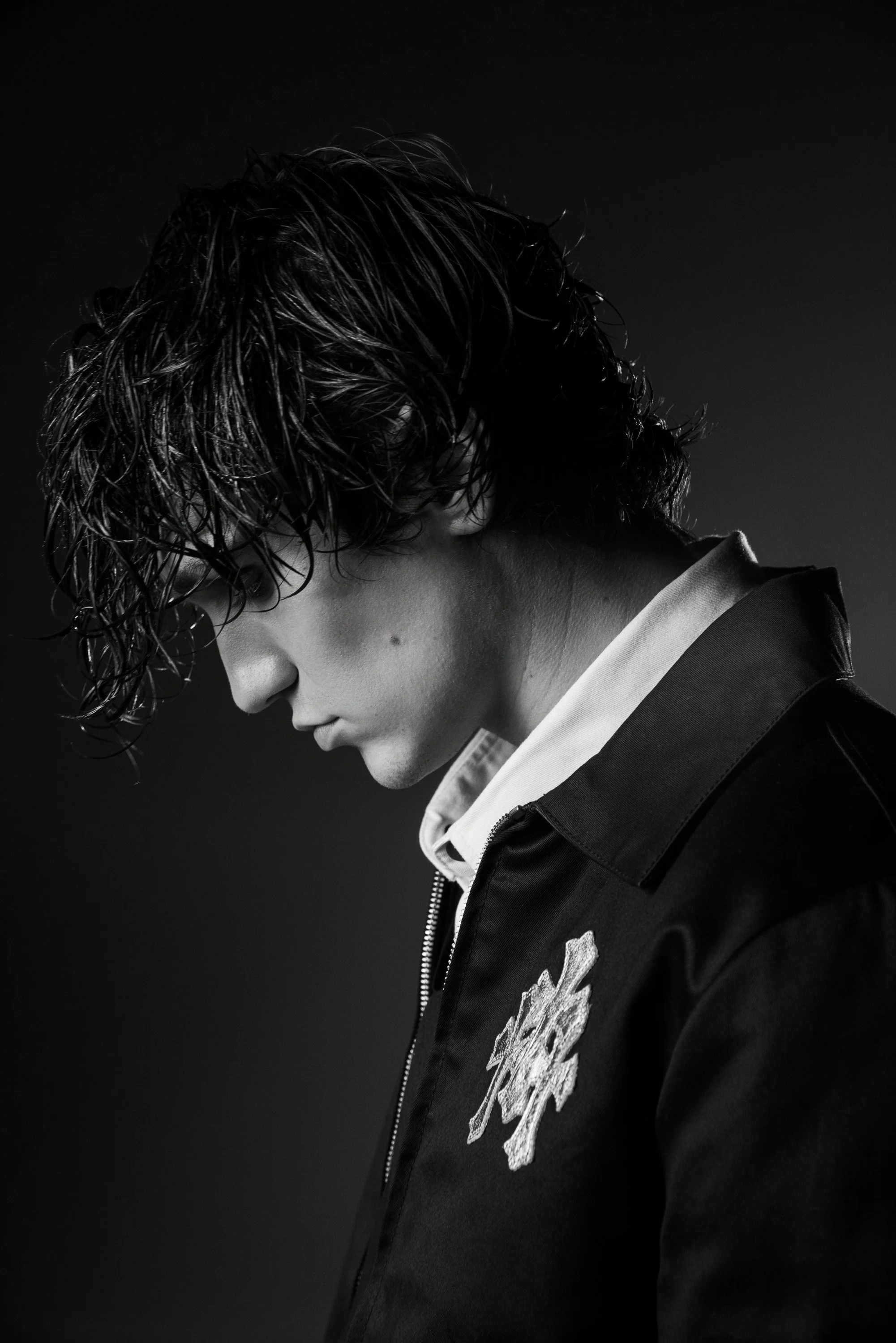 Black and white side profile of a young person with wet, wavy hair wearing a jacket with a floral patch.