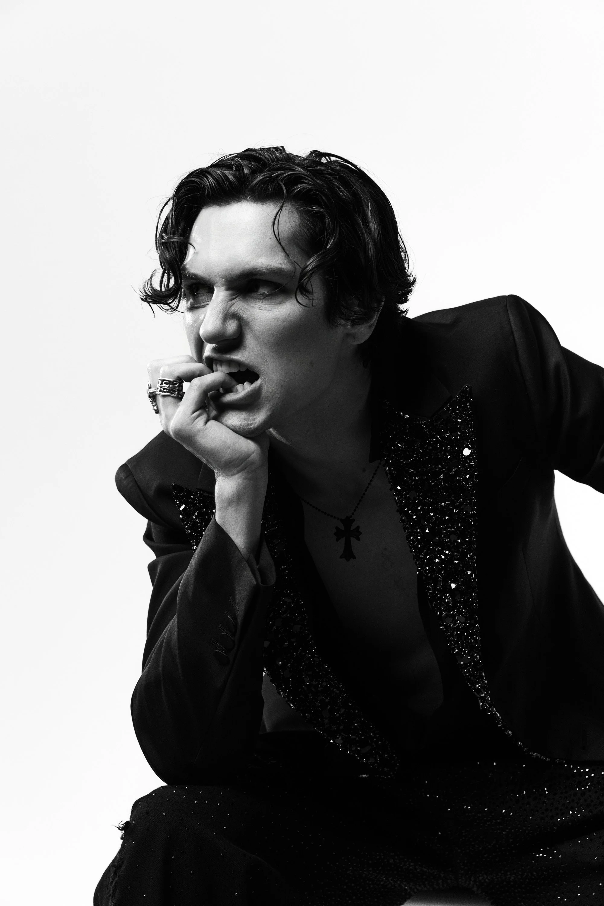 Black and white portrait of a young man with dark, wavy hair, wearing a black jacket with sequin details, a necklace with a cross, and rings, posing with his hand near his mouth against a plain white background.