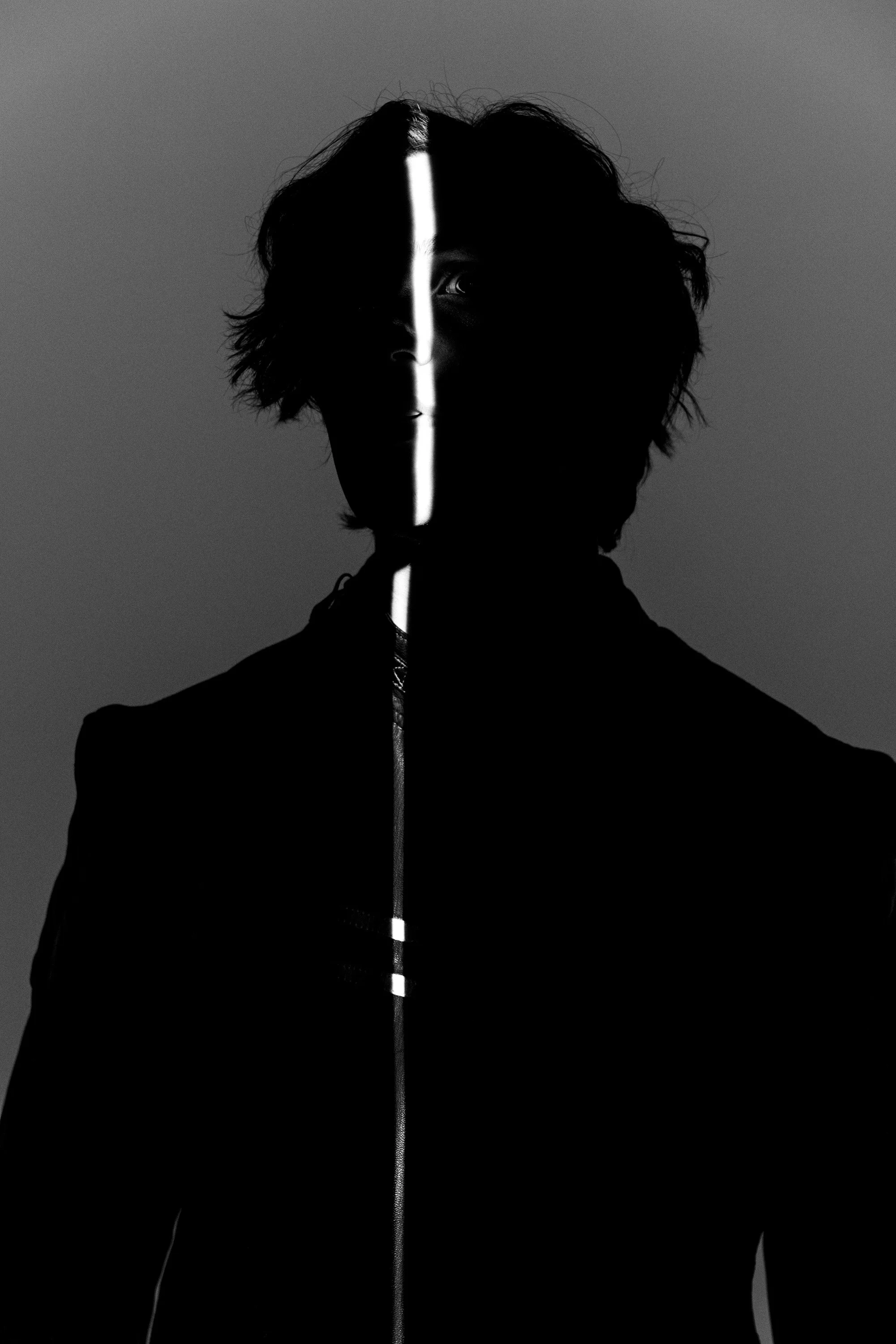 A black and white photo of a person with shoulder-length hair wearing a zippered jacket, with a vertical light stripe dividing their face, and the background is plain.