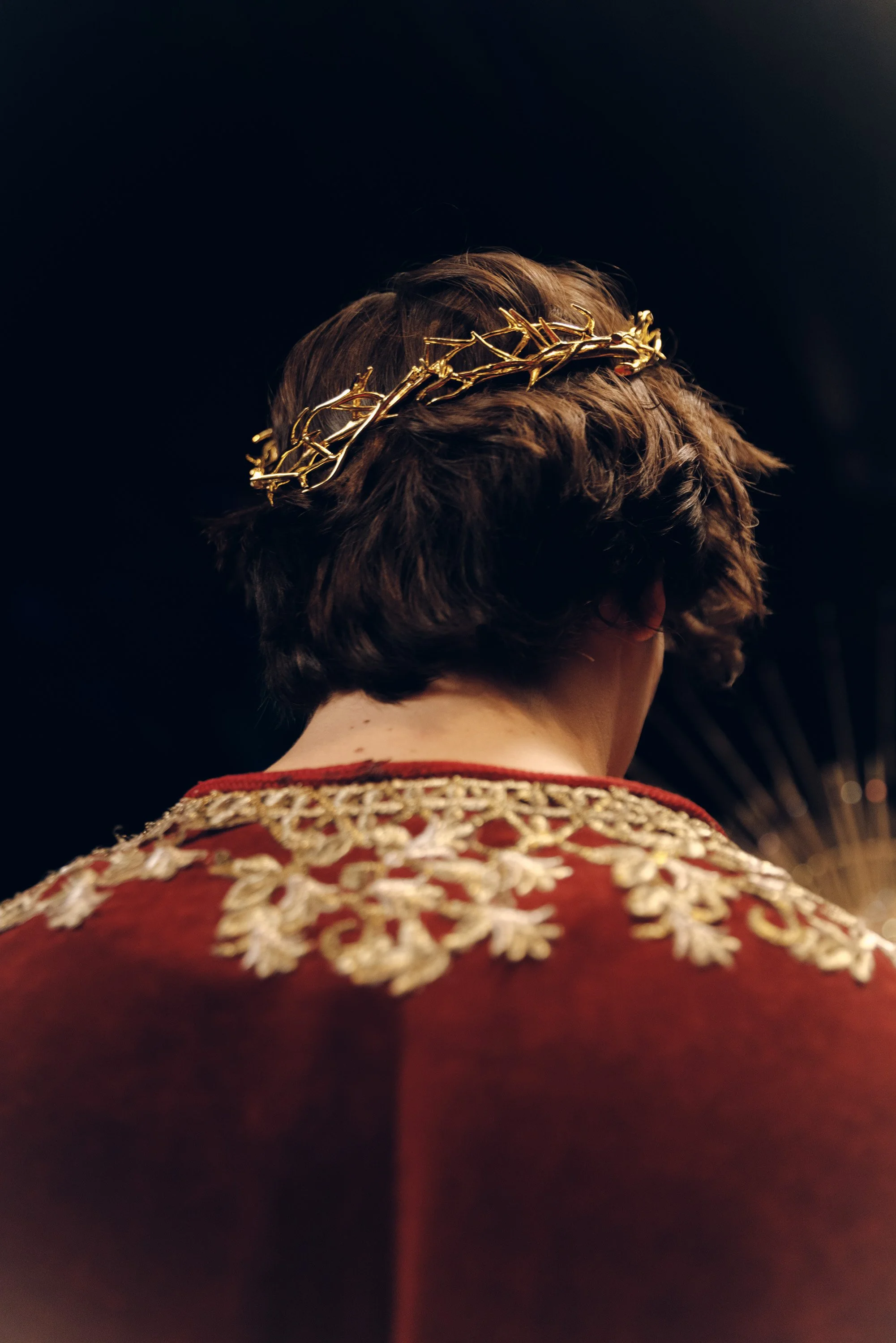 The back of a person's head with short, brown hair, wearing a gold-colored crown or headpiece. The person is dressed in a royal or formal red outfit with intricate gold embroidery on the shoulders. The background is dark, focusing on the person's hea