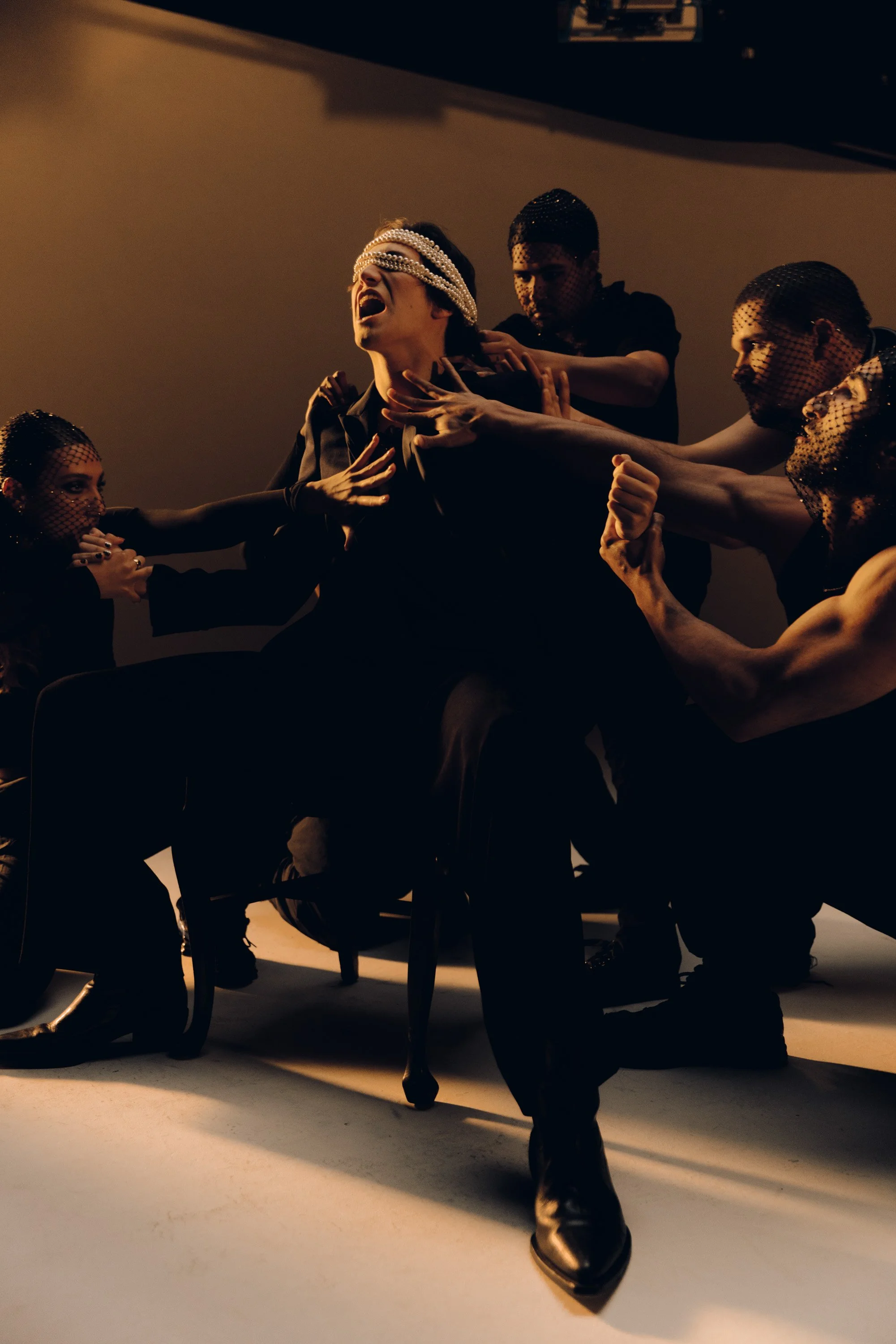 A group of five people with fishnet masks and black clothing in a dramatic pose, with one person sitting and wearing a pearl blindfold, and the others reaching towards them in a dark room with warm lighting.