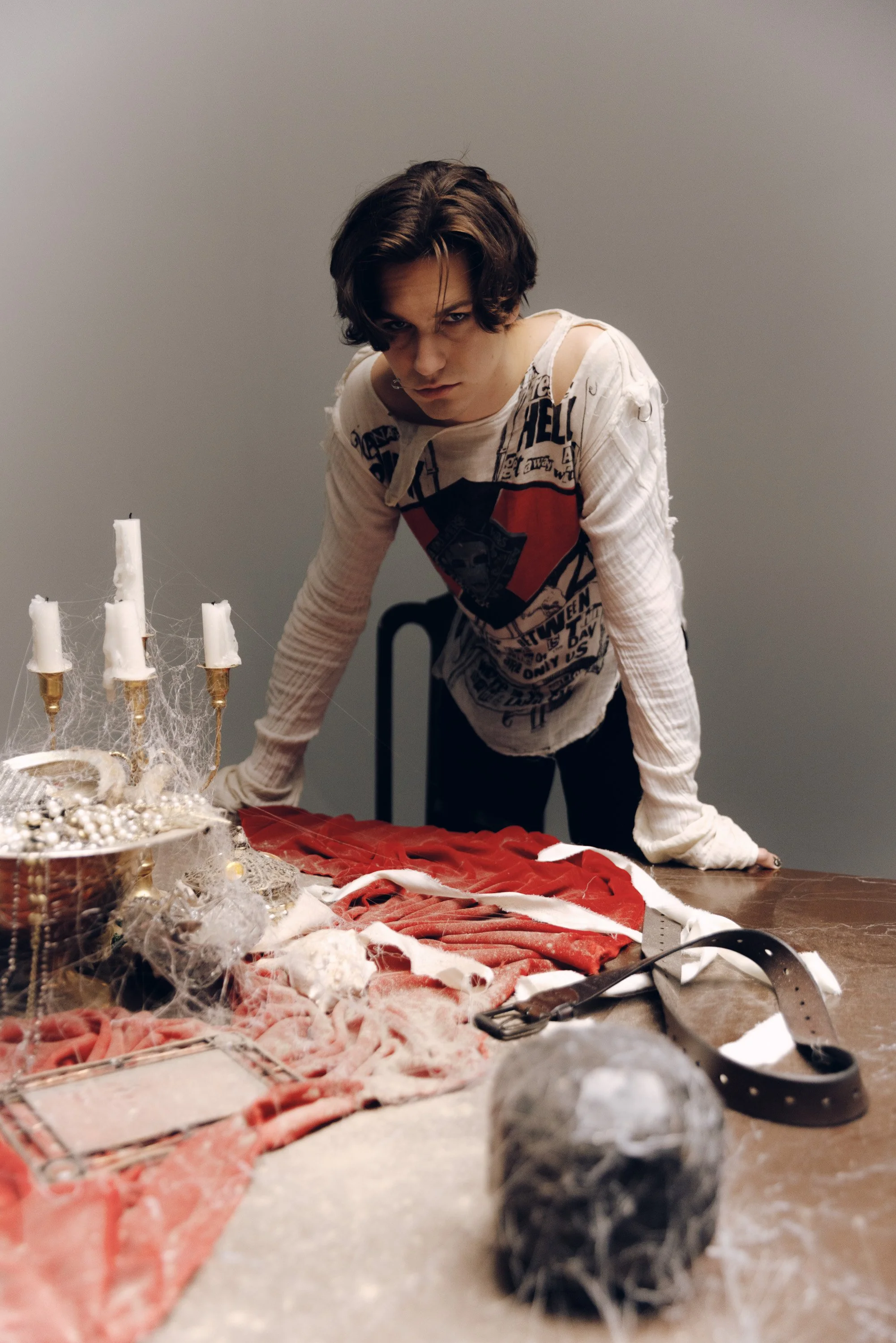 Person with dark, wavy hair wearing a white distressed long-sleeve shirt with a graphic design leaning over a table decorated for Christmas with a red tablecloth, candles, and ornaments, in front of a plain gray wall.