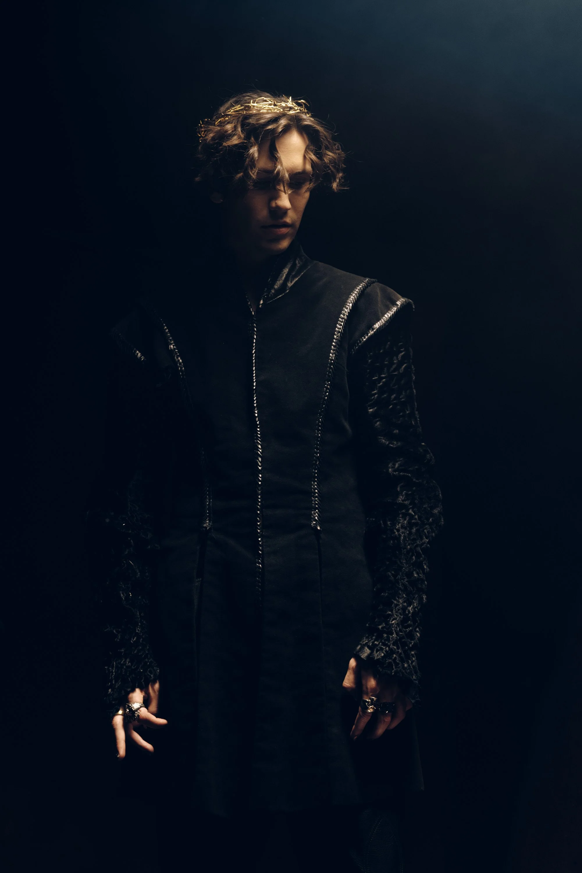 A person with curly hair wearing a crown and dark clothing, standing in a dimly lit environment.