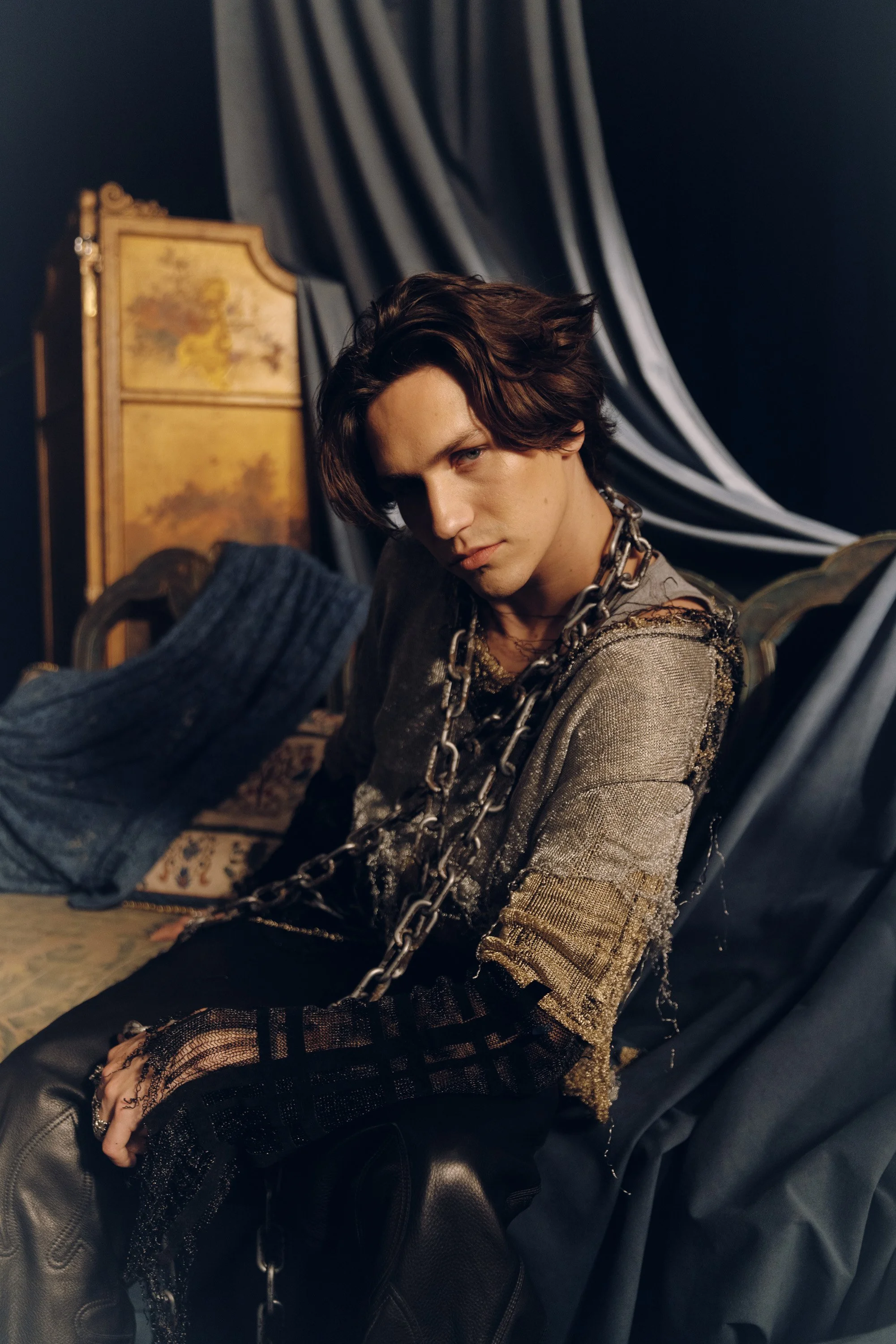 A young man with dark hair and a serious expression, wearing a metallic gold and black outfit, sitting on a black leather chair among ornate furniture and dark drapery, with chains draped over his shoulders.