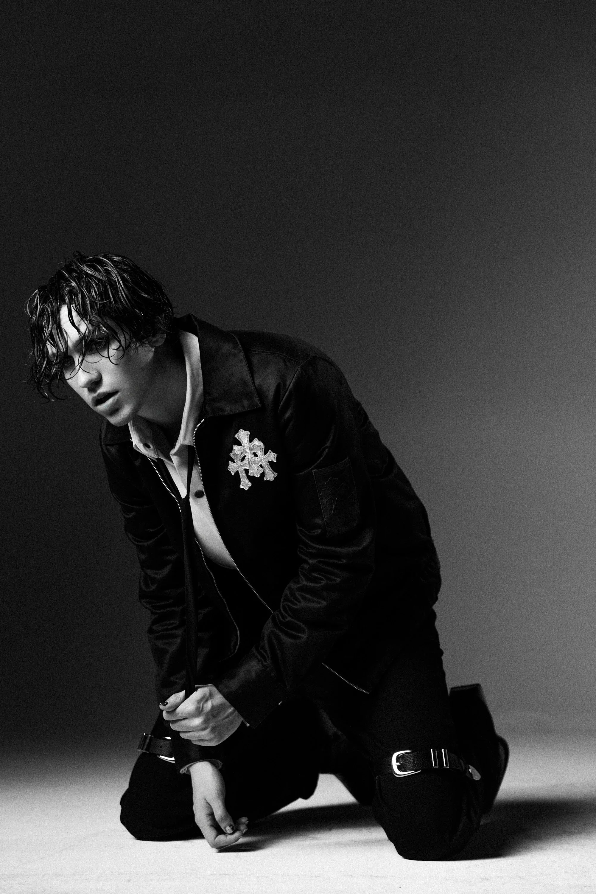 Black and white photo of a person with wet hair, wearing a jacket with a snowflake emblem, kneeling on the floor with one hand on the ground and the other hand holding their wrist.