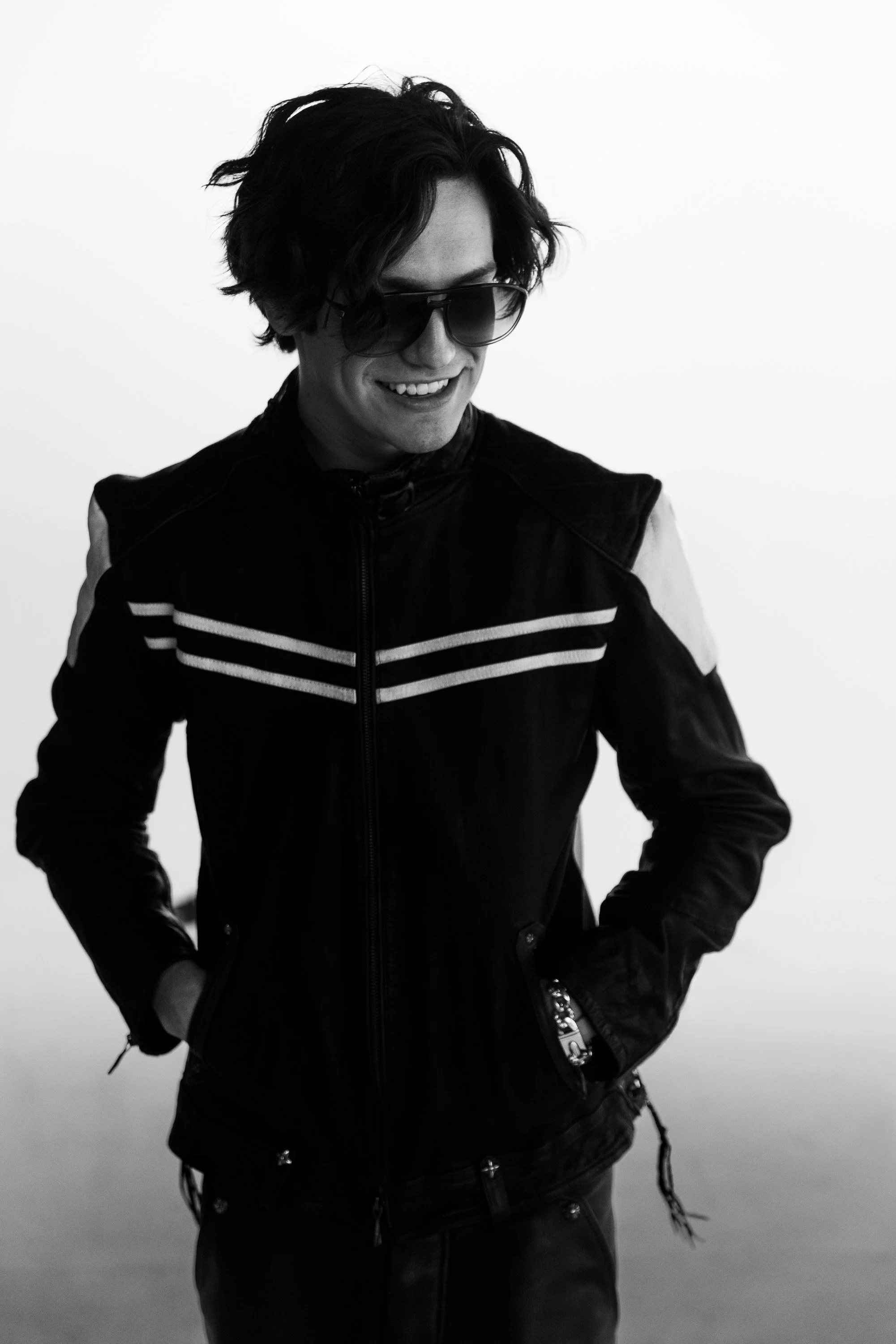 A young man with wavy dark hair wearing sunglasses and a black leather jacket, smiling and standing against a plain background.