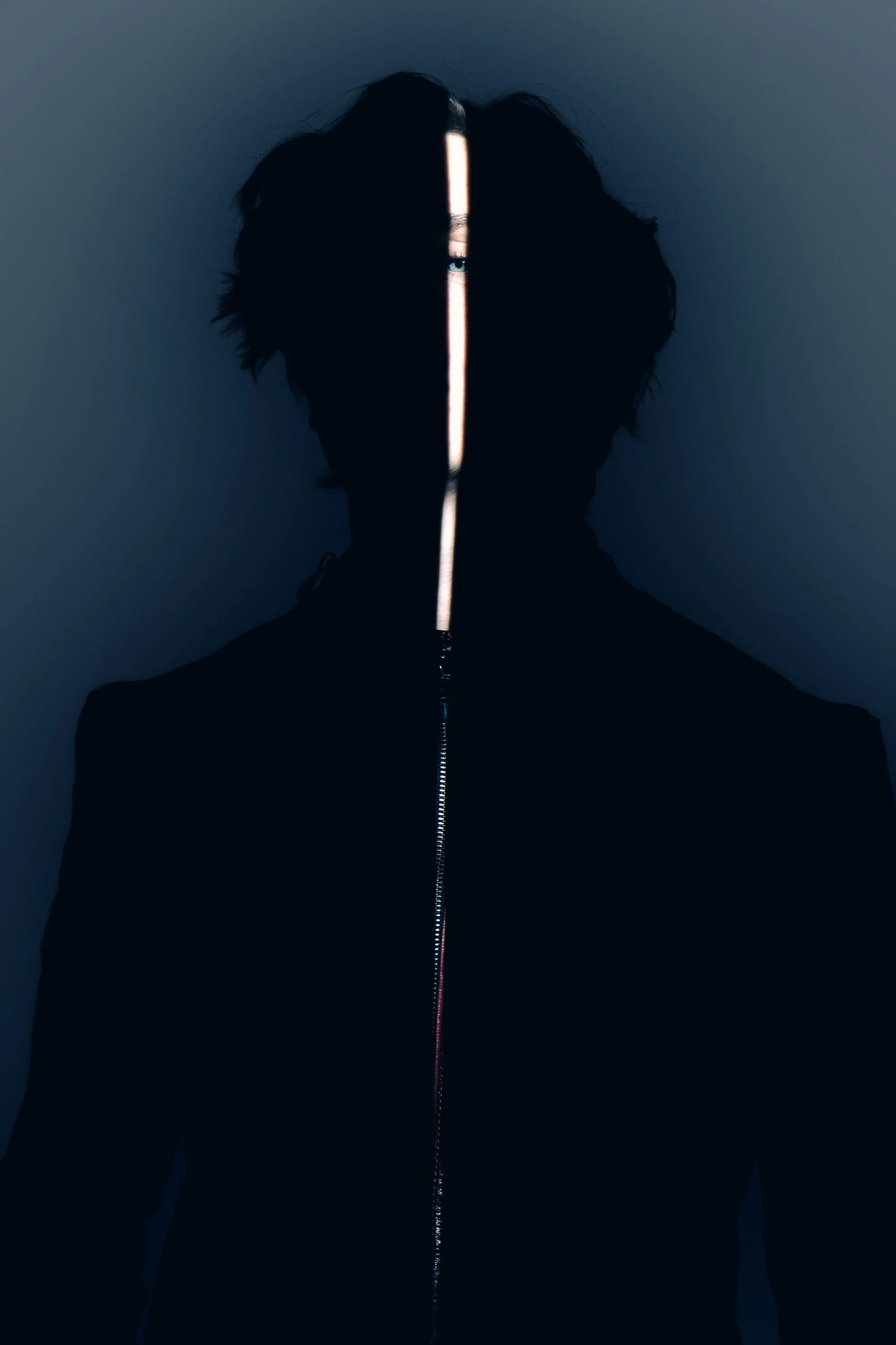 A person with curly dark hair wearing a black zip-up hoodie, partially obscuring their face with a vertical light or object in front of it, set against a plain dark background.