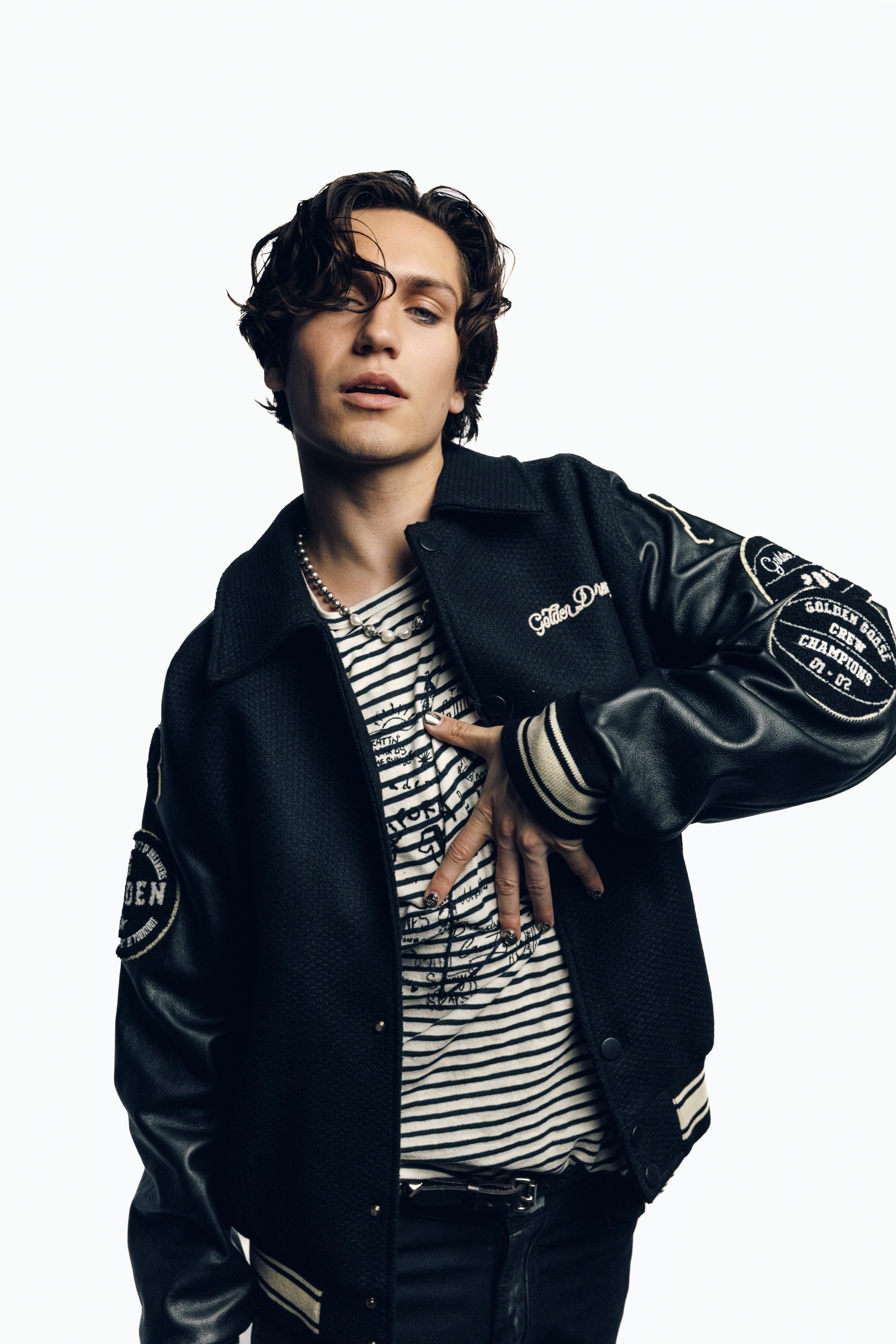 A young man with curly dark hair and a confident expression, dressed in a black varsity jacket with patches, a striped shirt, and layered necklaces, posing with one hand on his chest against a plain white background.