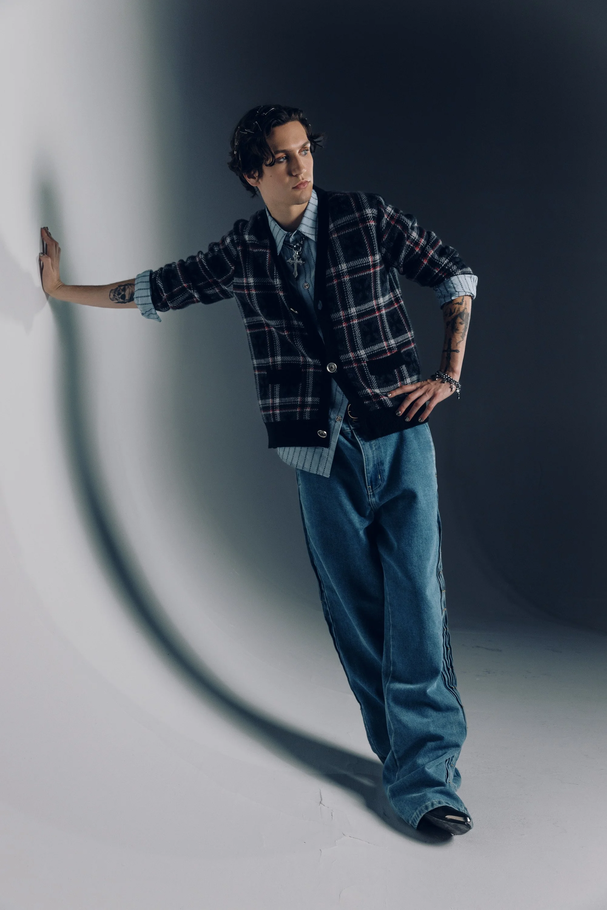 A young man with dark wavy hair, tattoos on his left arm, wearing a plaid jacket over a striped shirt, denim jeans, and black shoes, poses against a two-toned background, leaning with his right hand on a wall and looking away from the camera.