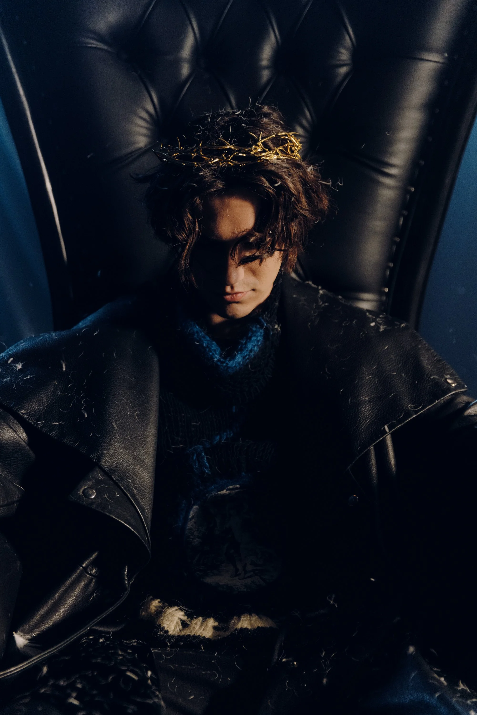 A person with curly hair and a gold crown sitting on a black leather chair, wearing a black fur coat, with their head slightly bowed and eyes closed.