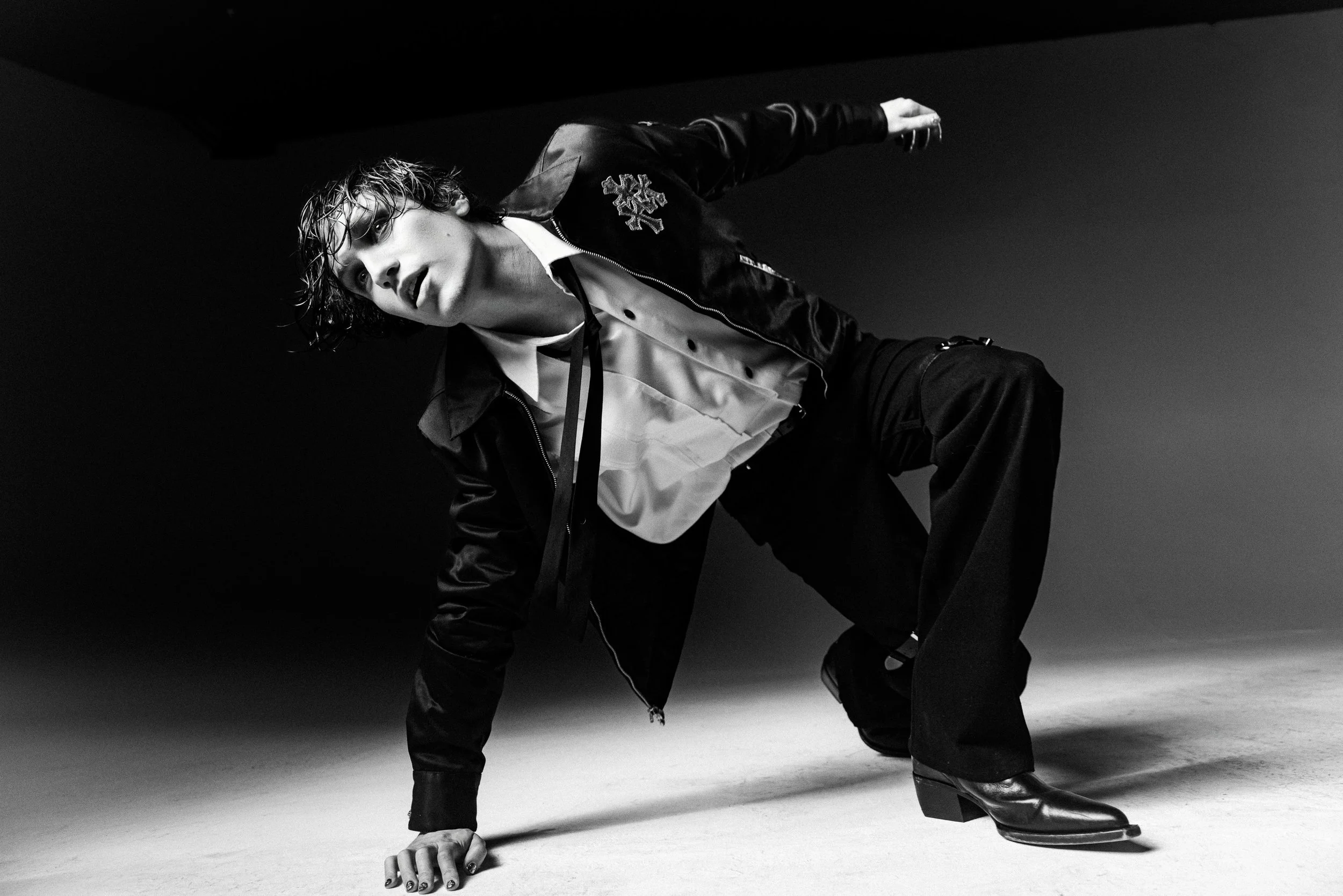 Black and white photo of a woman with wet hair, wearing a jacket with patch details, leaning on the ground with one hand and one knee, in a studio setting.
