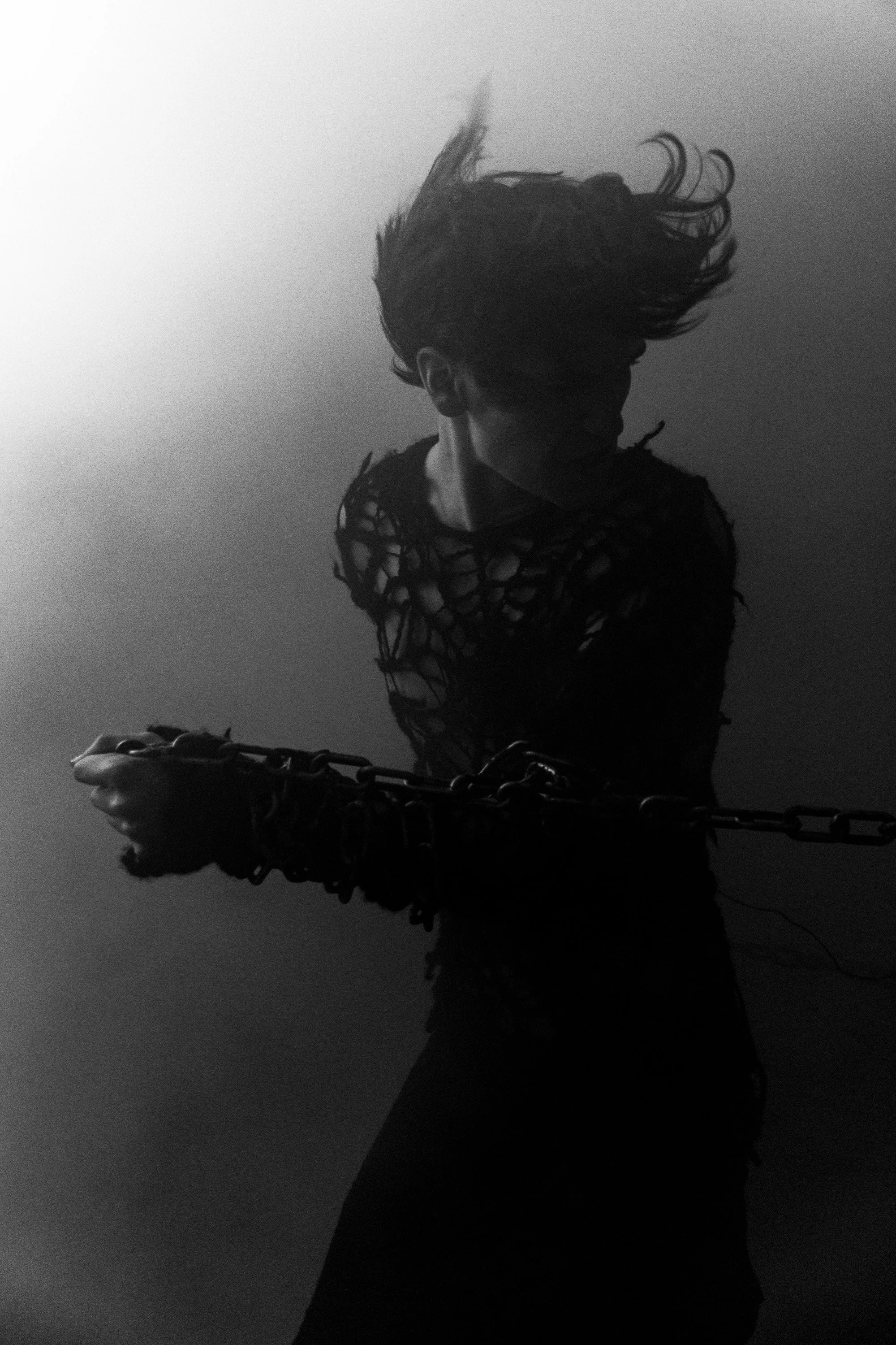 A person with long, flowing hair, wearing a torn, mesh-like black outfit, holding chains, standing in front of a plain, gradient background.