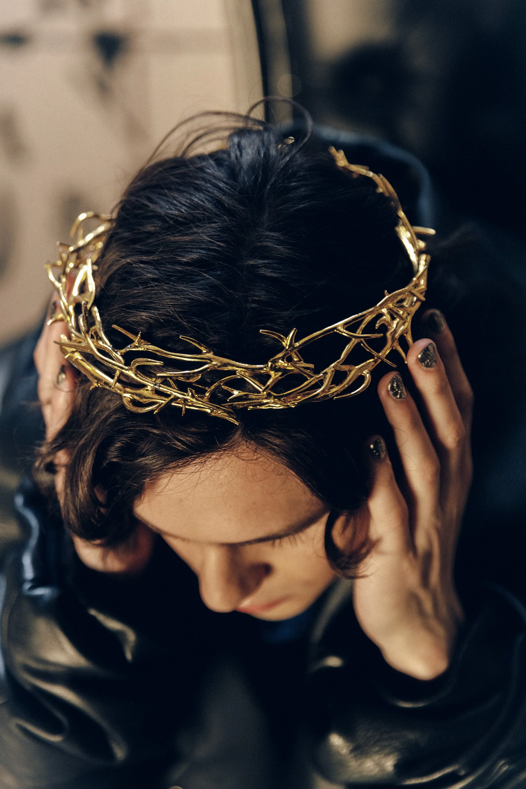 Top-down view of a woman with dark, curly hair wearing a golden crown with intricate thorn-like design, holding her head with her hands, nails painted with black and gold glitter, near a black leather surface.