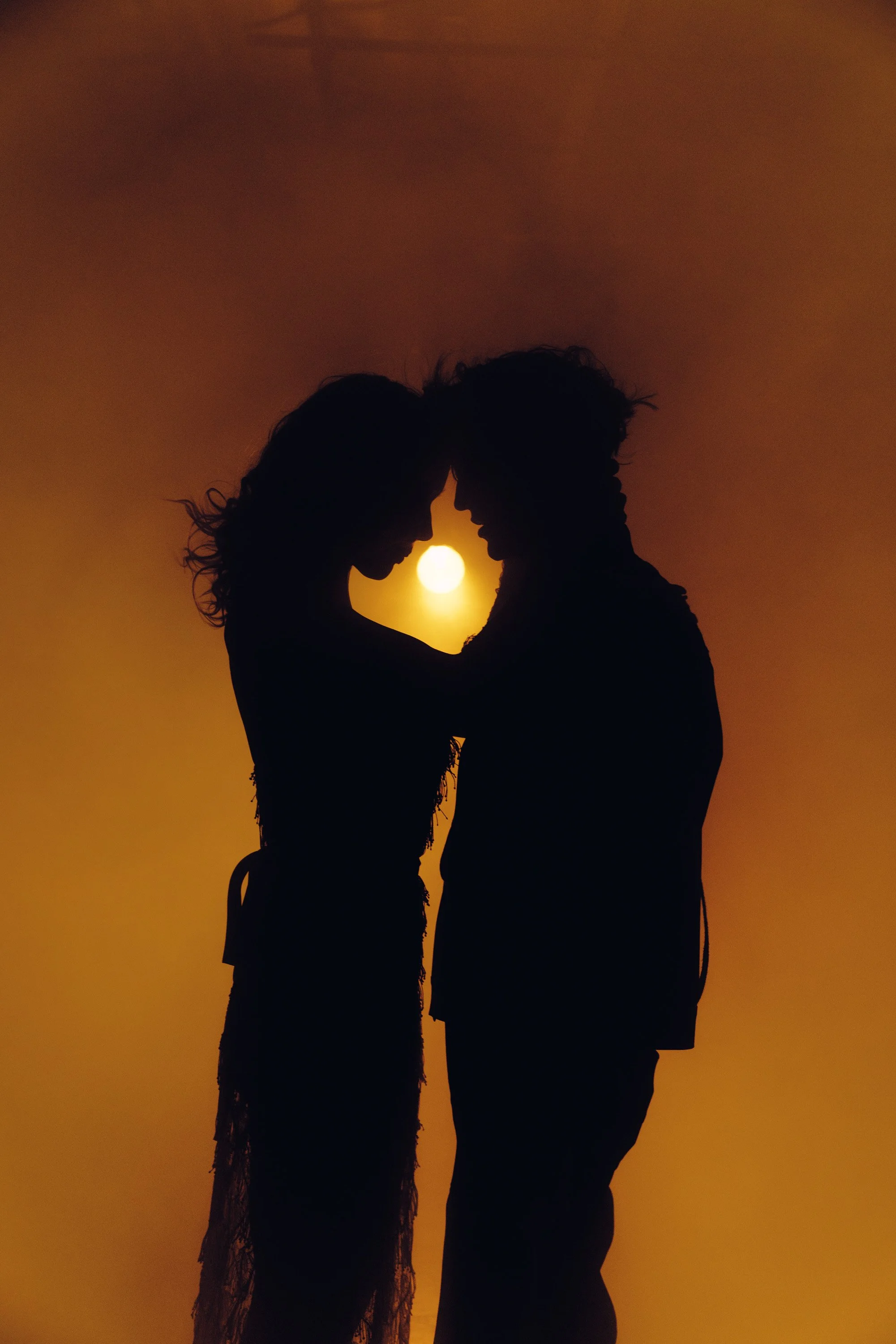 Silhouettes of a couple facing each other with their foreheads touching, forming a heart shape with their arms, against a sunset with the sun centered between them.