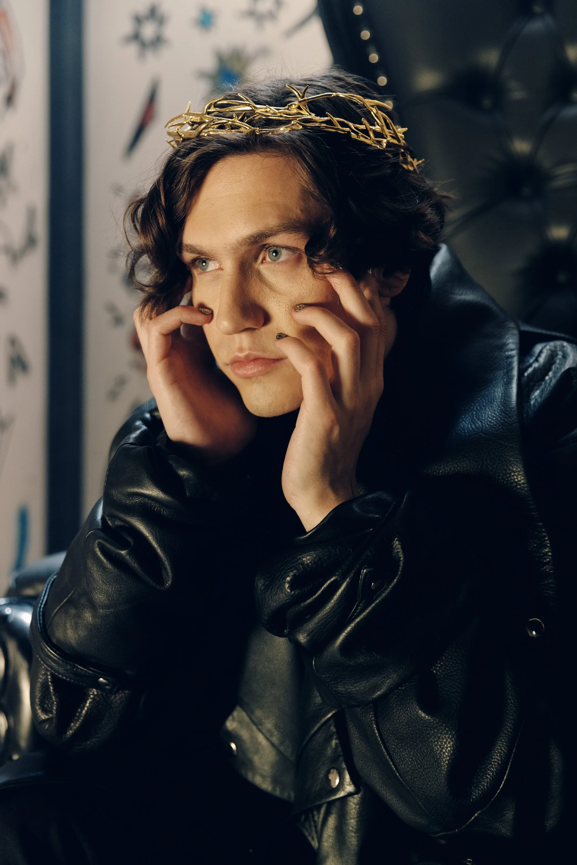 A person with shoulder-length dark hair wearing a gold crown, a black leather jacket, and gray shirt, resting their face on their hands in front of a decorated wall with snowflakes and star shapes.