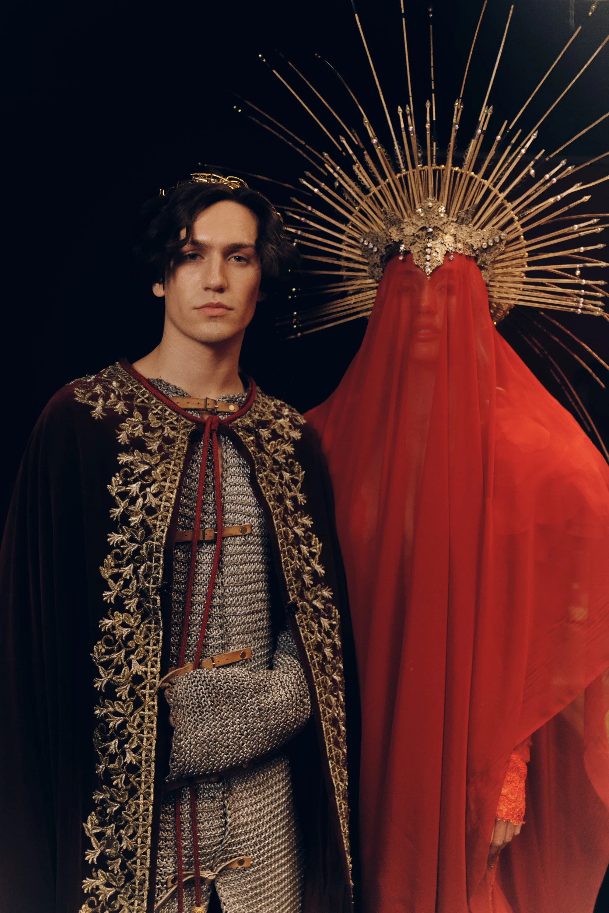 A young man with dark wavy hair dressed in an ornate, embroidered robe stands next to a religious statue with a golden headdress, veil, and flowing red fabric, both set against a dark background.