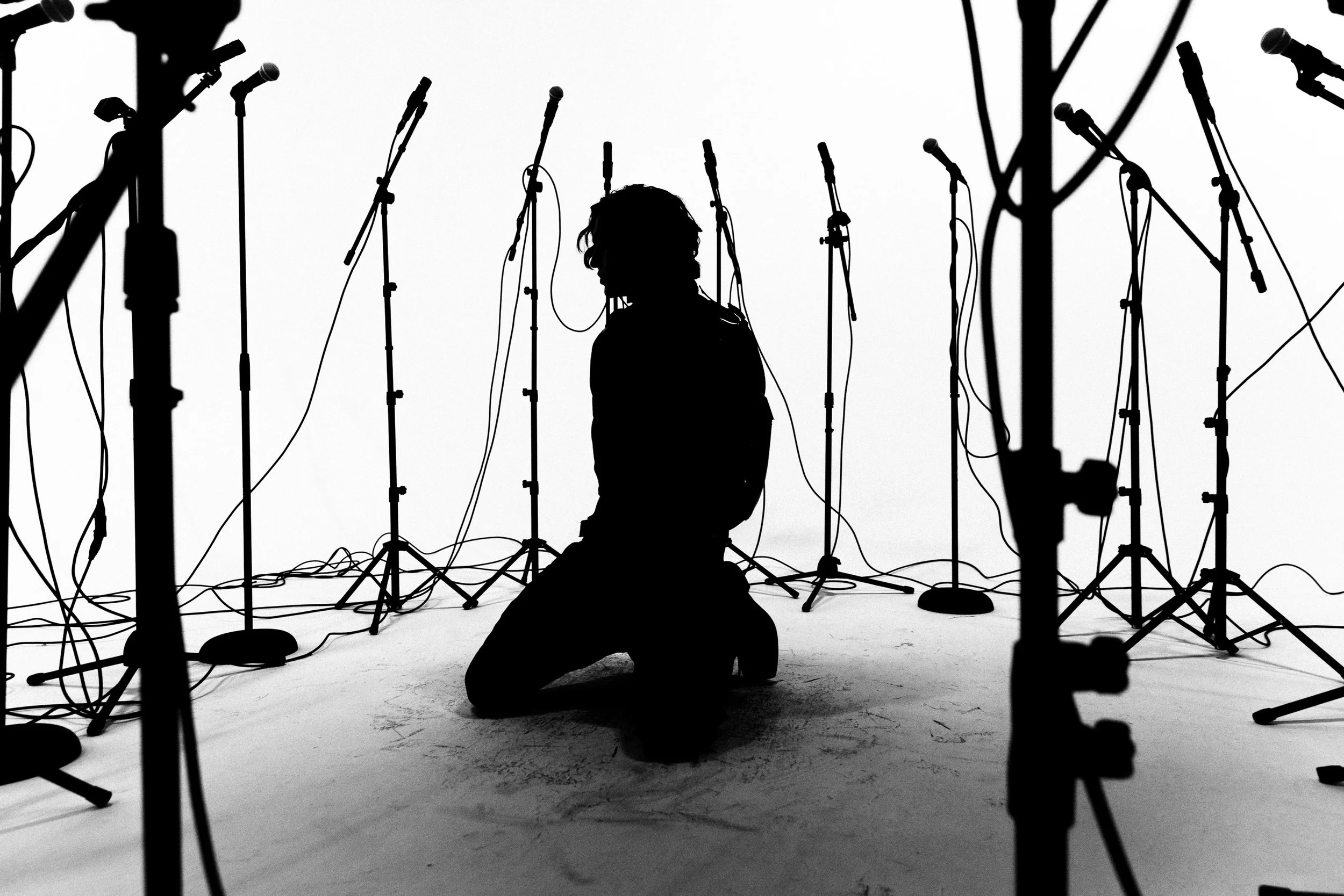 Silhouette of a person kneeling among multiple microphone stands in a recording studio or stage setup.