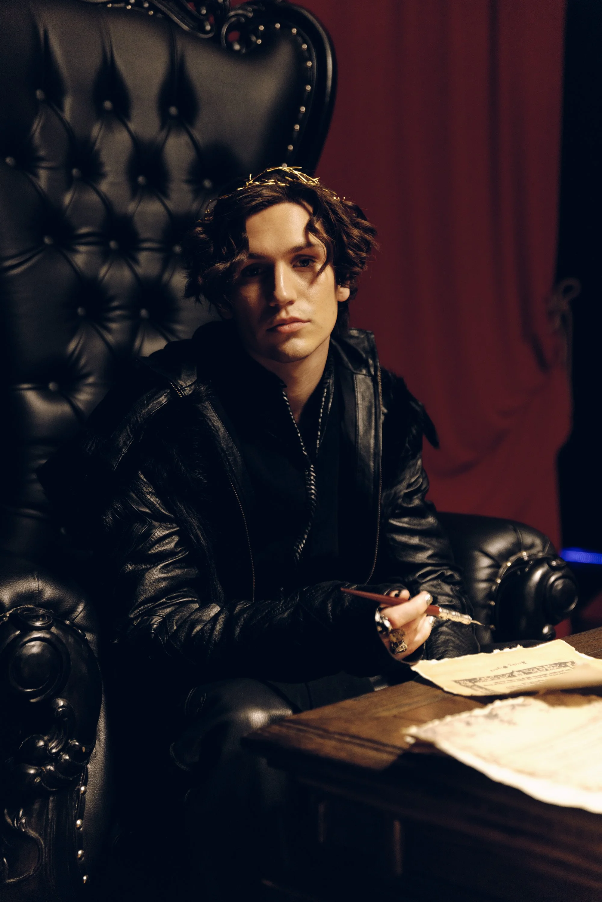 A young woman with dark wavy hair, wearing a crown and leather jacket, sitting on a black tufted leather armchair in a dimly lit room, holding a pen over papers on a wooden table.