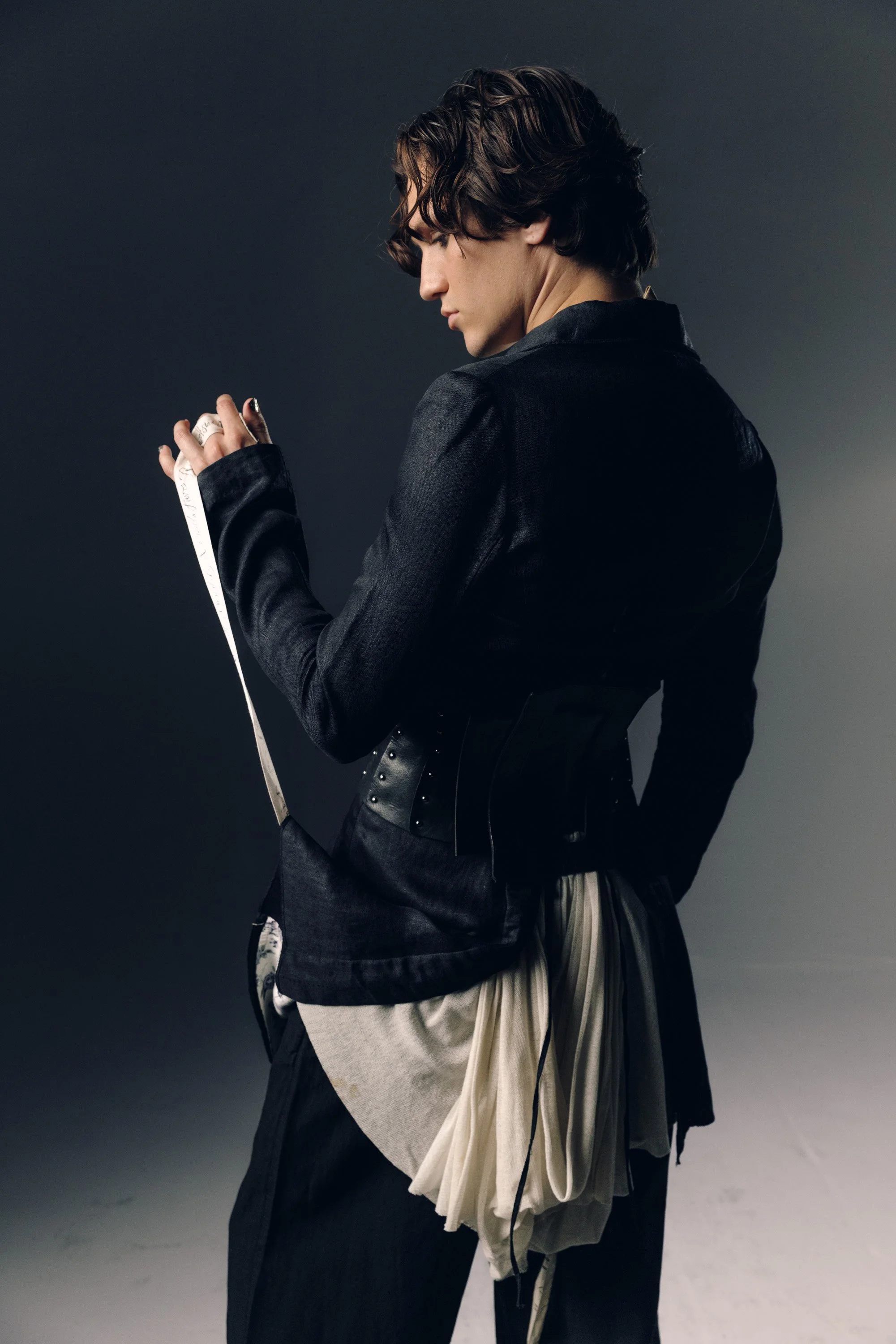 A person dressed in black clothing, looking down while holding a measuring tape with both hands, standing against a gray background.