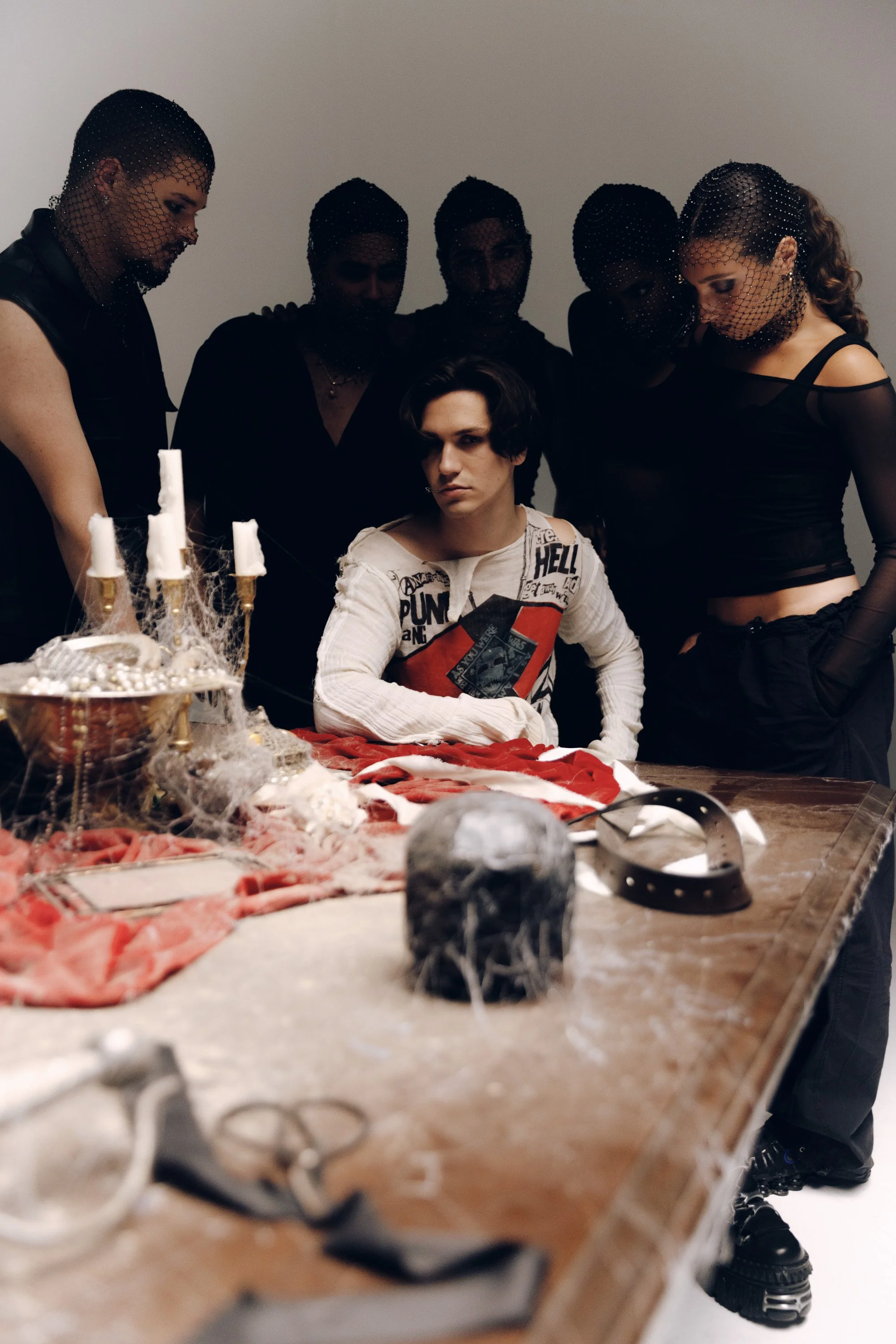 A group of people gathered around a table decorated for Halloween with cobwebs, candles, and a skull. One person sits at the table with an intense expression, while the others stand behind and around him, some wearing black clothing and fishnet masks
