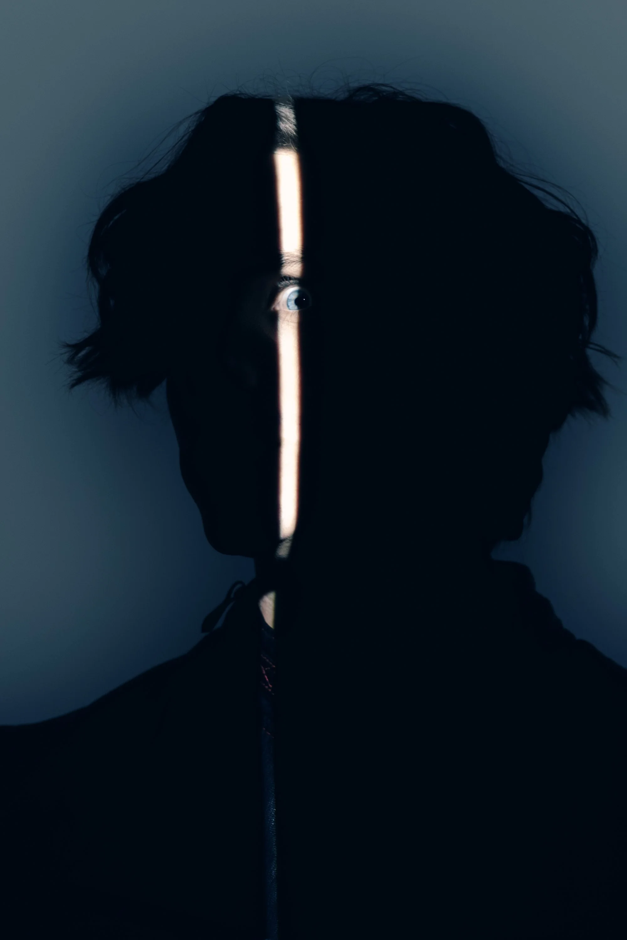 A person in dark clothing with curly hair features a narrow vertical strip of light illuminating their face, showing one blue eye, while the rest of the face is in shadow.