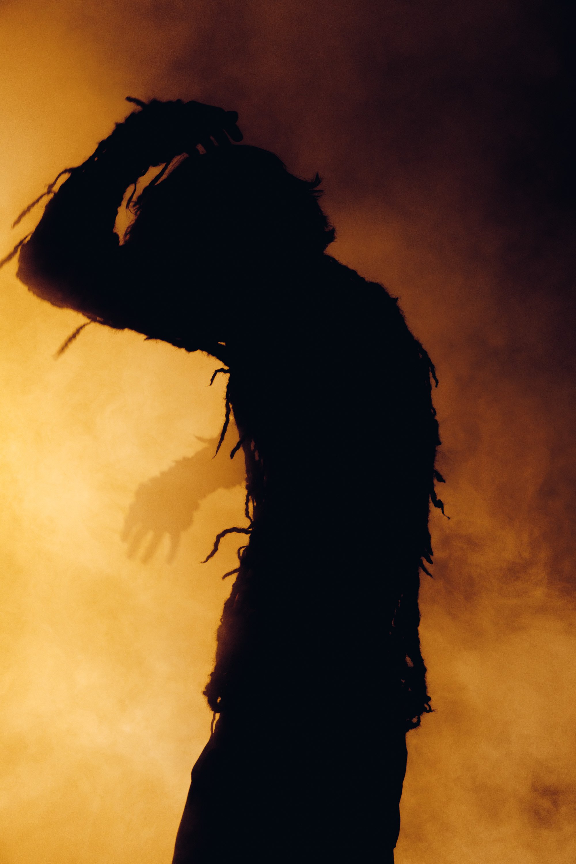 Silhouette of a person with dreadlocks, holding their head with one hand, against a warm, gradient background with orange and brown tones.
