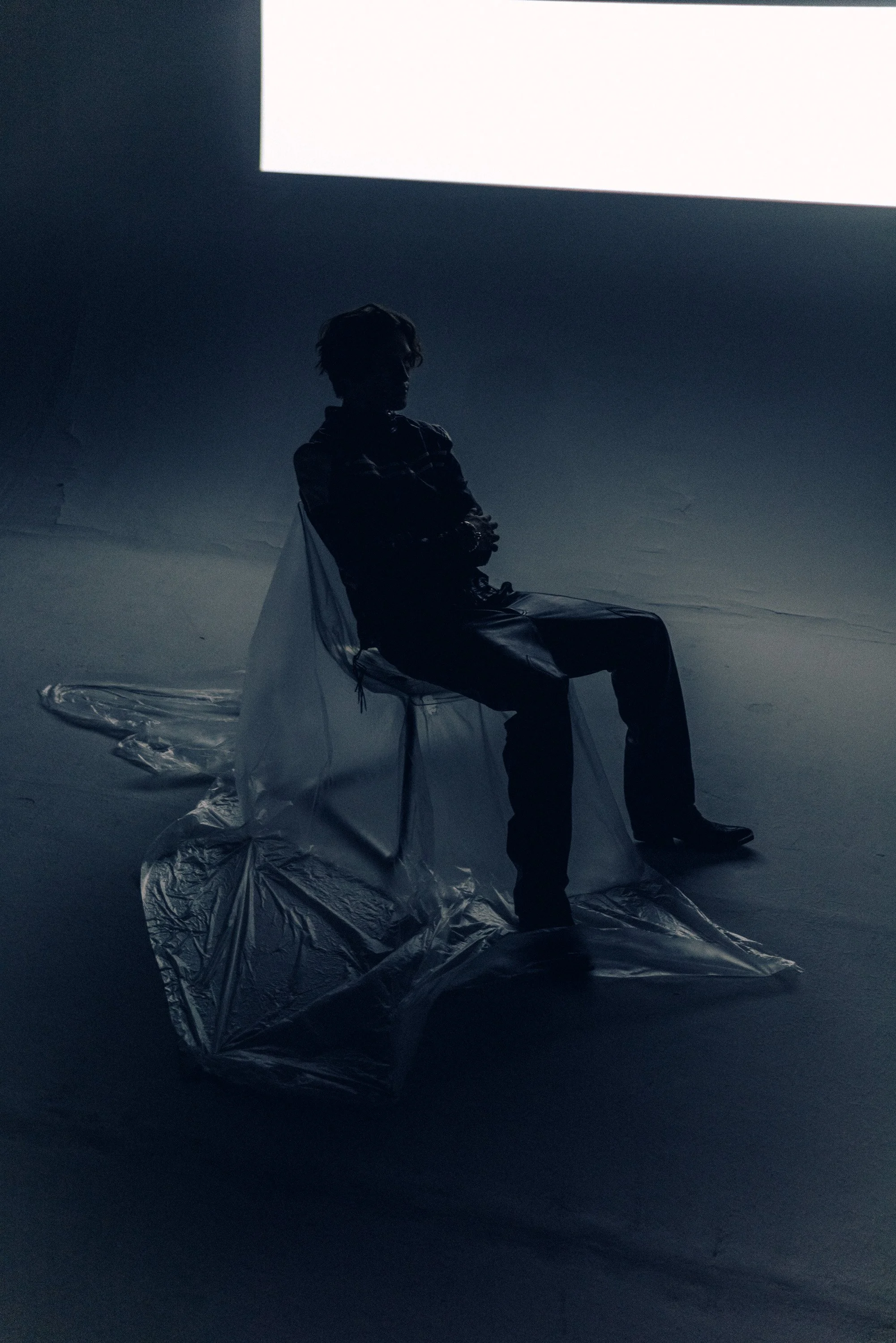 A person sitting on a transparent plastic sheet in a dark room, illuminated by a bright white screen or light source above.
