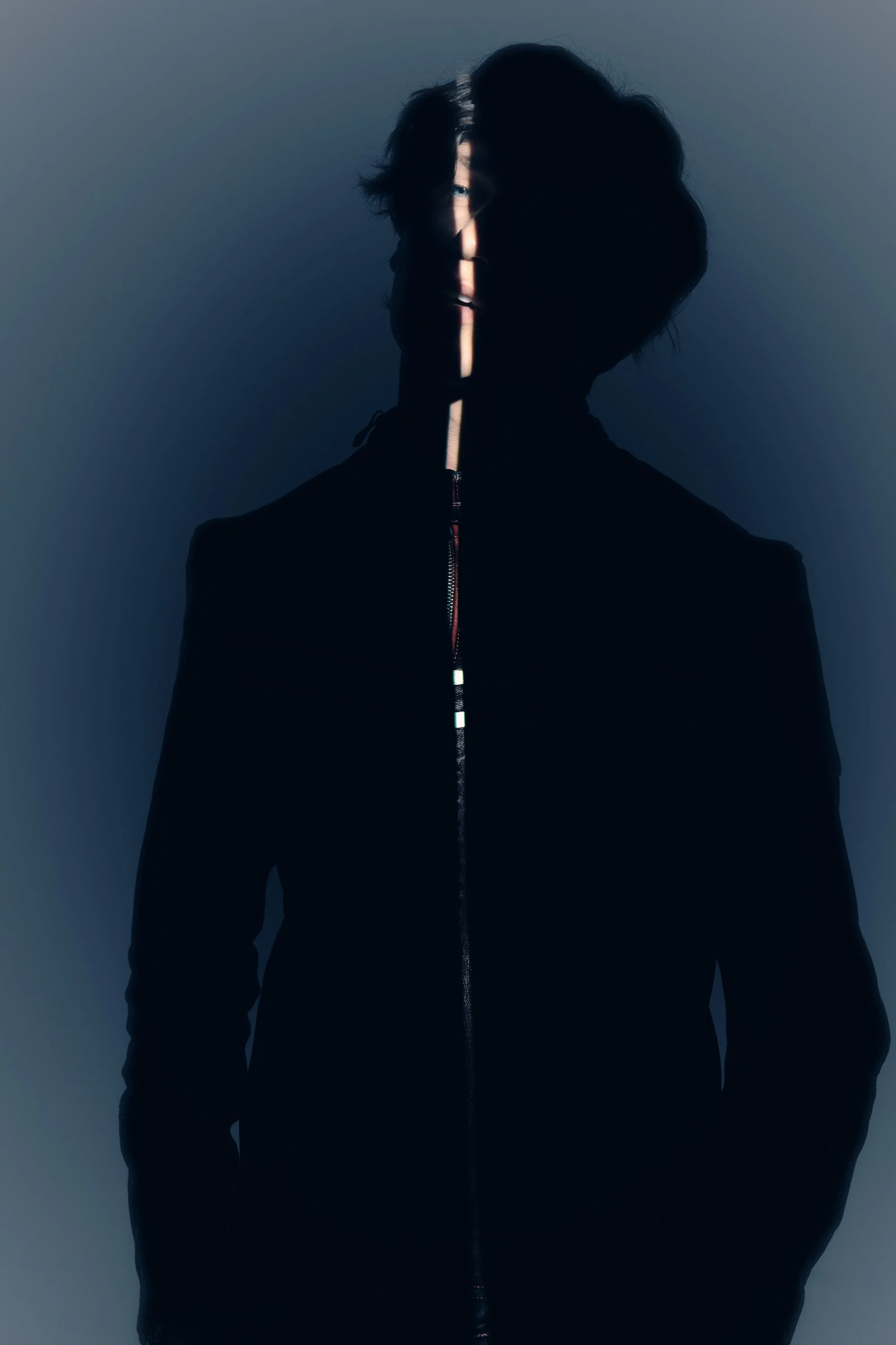 A person with dark, wavy hair wearing a black zip-up jacket, standing against a plain background with light and shadow creating a partially obscured face.
