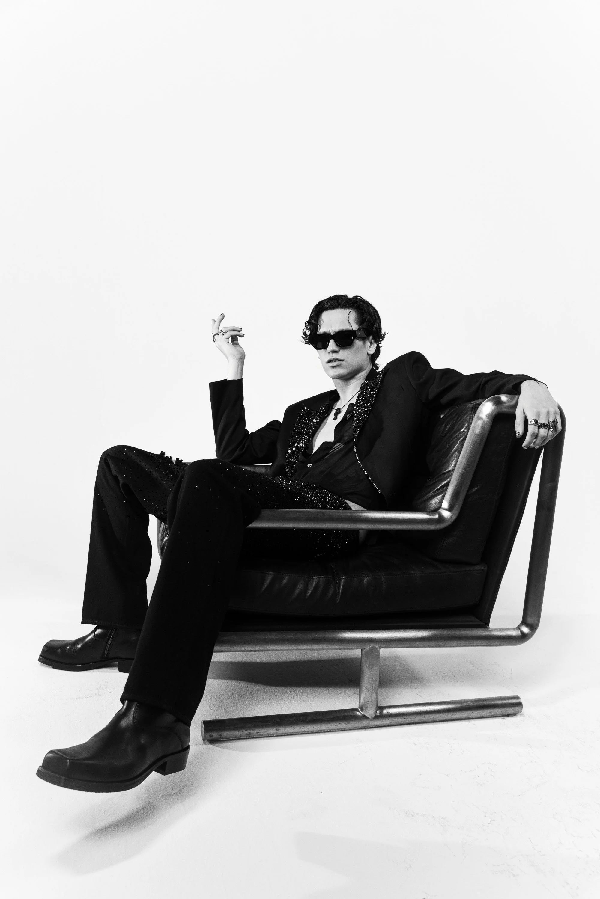 Black and white photo of a young man with wavy hair wearing sunglasses, a dark blazer with glittering details, and platform boots, sitting on a modern chair against a plain white background.