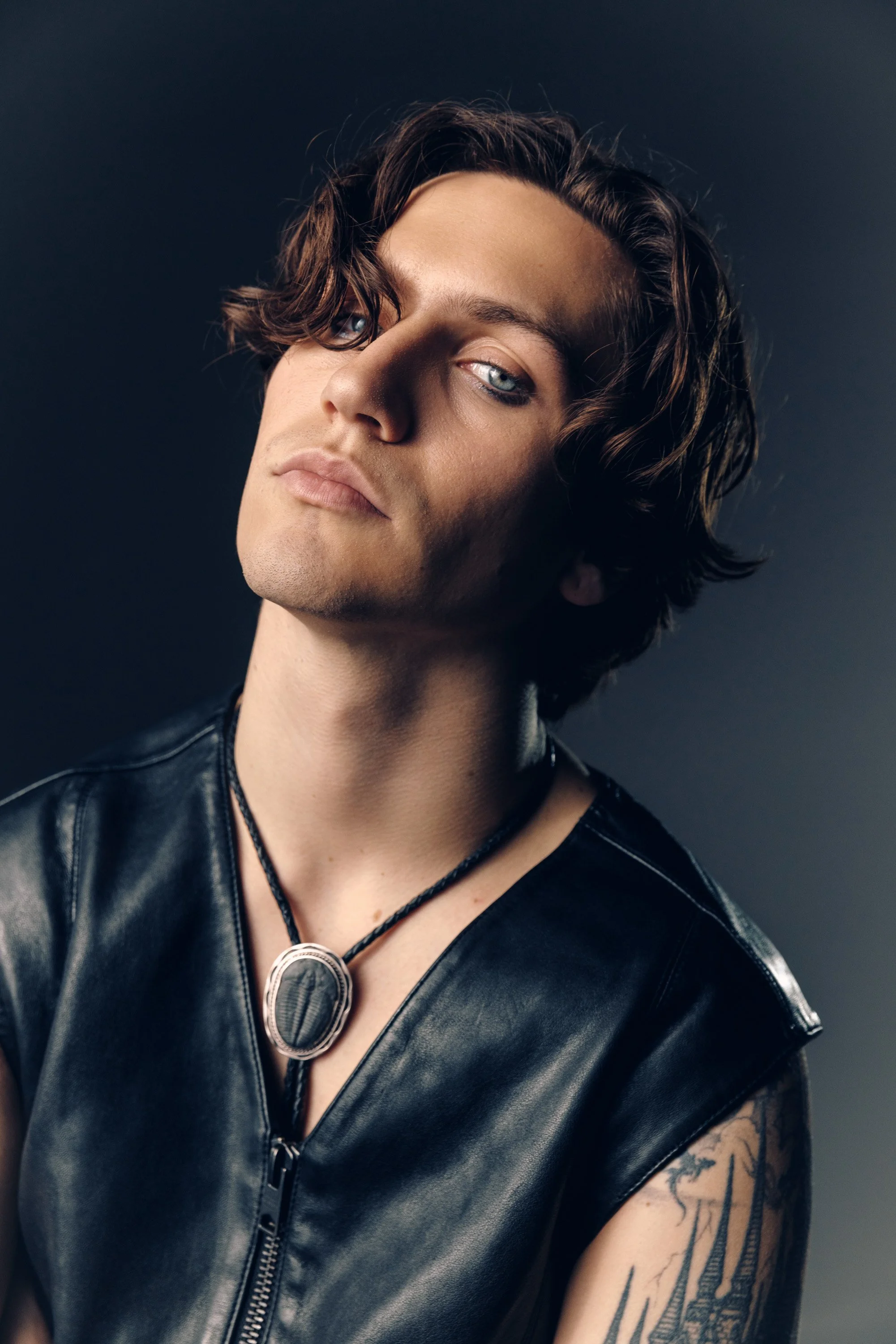 A young man with shoulder-length wavy brown hair, piercing blue eyes, and a light complexion. He is wearing a black leather vest with a zipper, a black necklace, and sunglasses hanging from his shirt. He has visible tattoos on his left arm and is pos