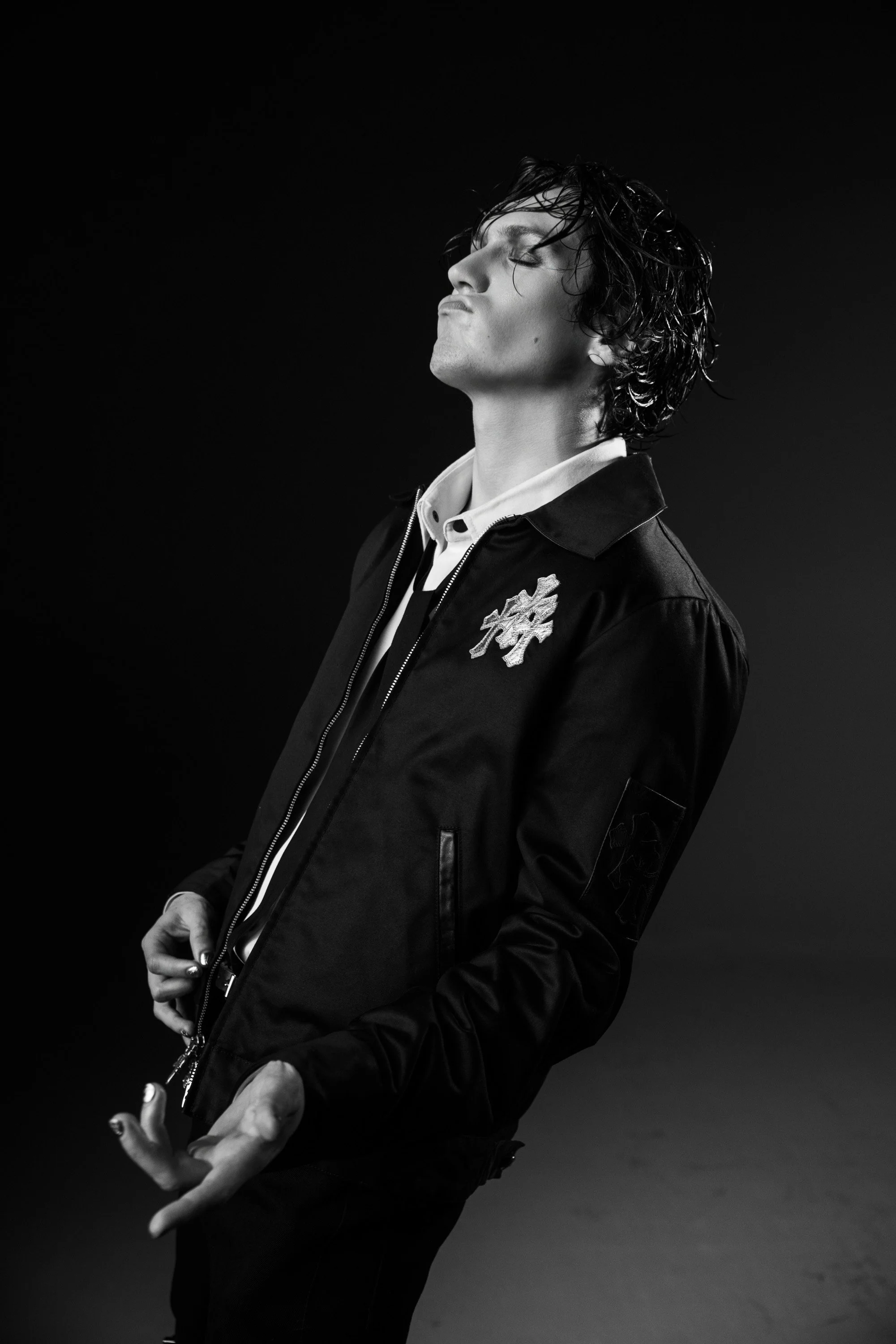 Black and white photo of a young man with wet hair, eyes closed, wearing a jacket with a patch, slightly tilting his head back with hands positioned in front of him.