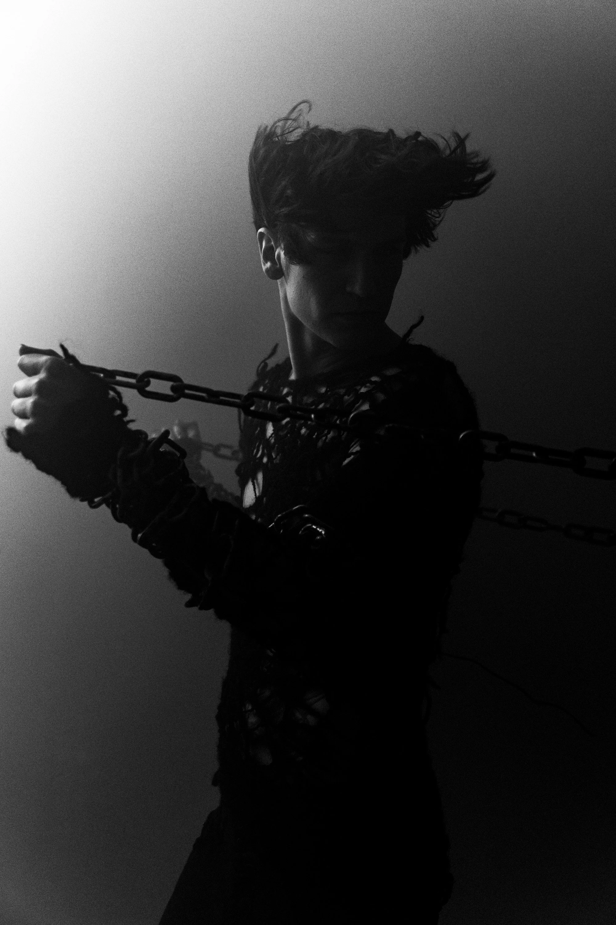 A person with curly hair holding a chain in a dark setting, with a high contrast black and white filter.