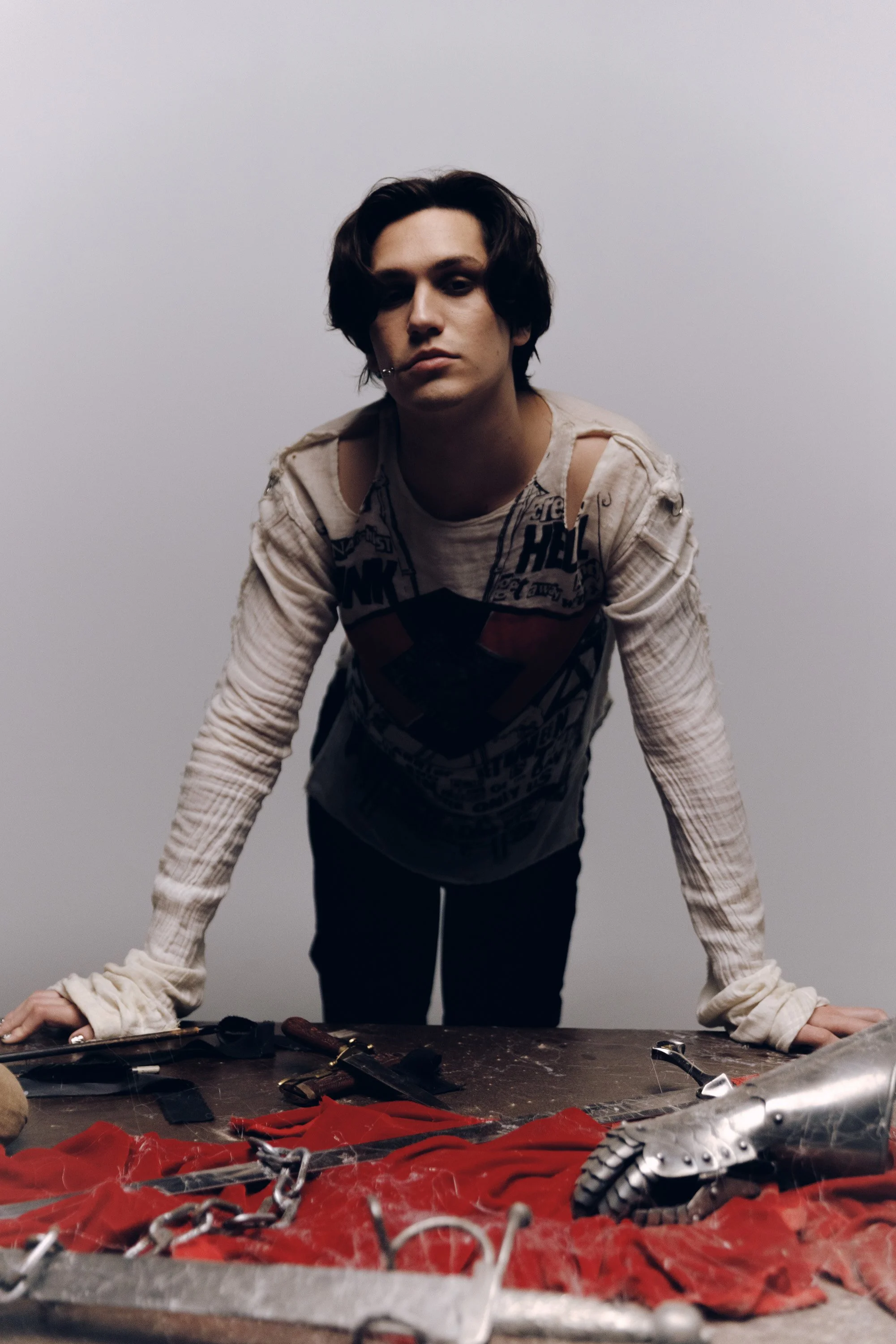 A person with dark, wavy hair and a piercing on the lower lip leans over a cluttered worktable, with a blank, light gray background. The table has various tools, chains, red fabric, and broken metal parts.