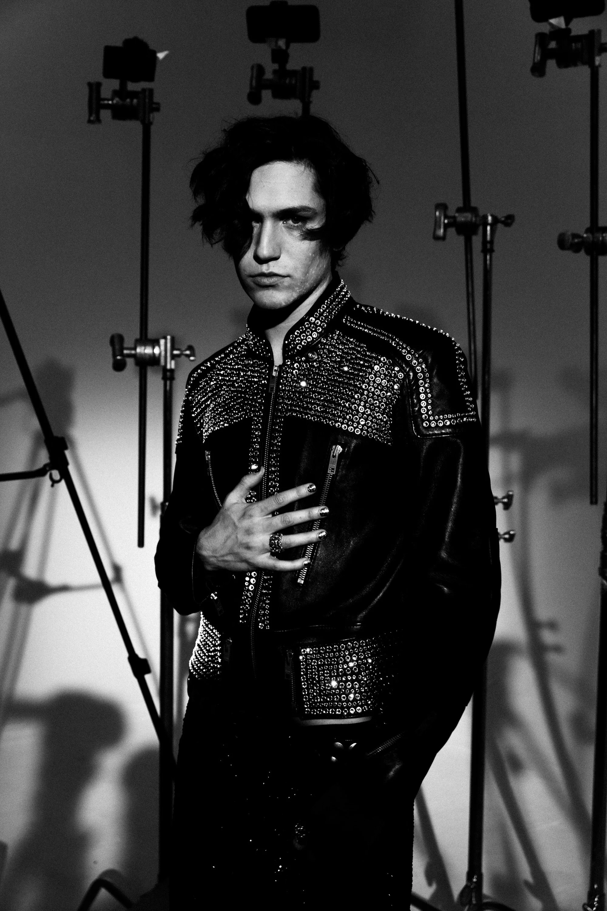 A person with dark, wavy hair and light skin posing with their hand on their chest, wearing a jacket decorated with rhinestones or studs, surrounded by studio lighting equipment in a black-and-white photograph.