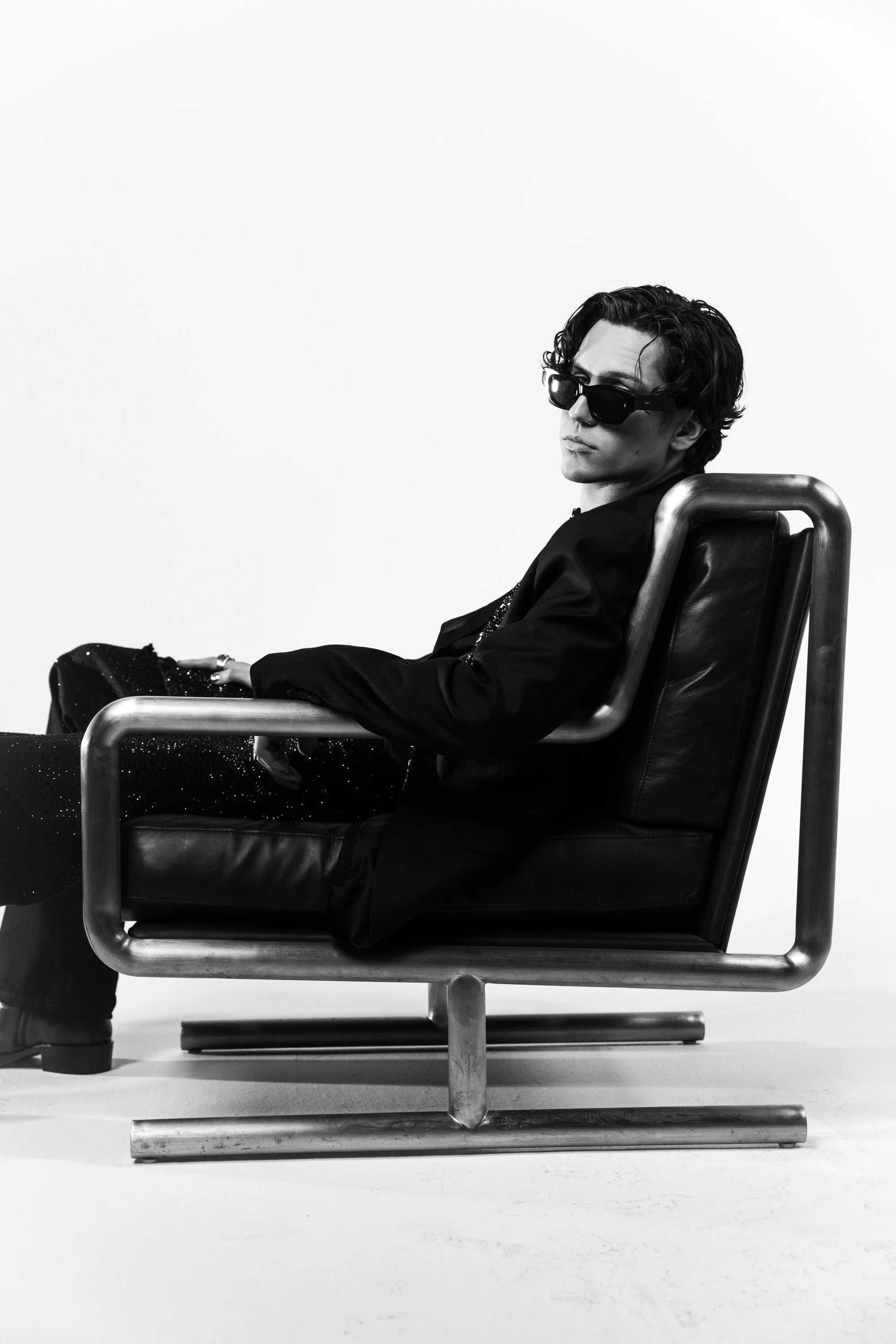 A black and white photograph of a young man sitting in a modern chair, wearing sunglasses and a dark outfit, with a serious expression, against a plain white background.