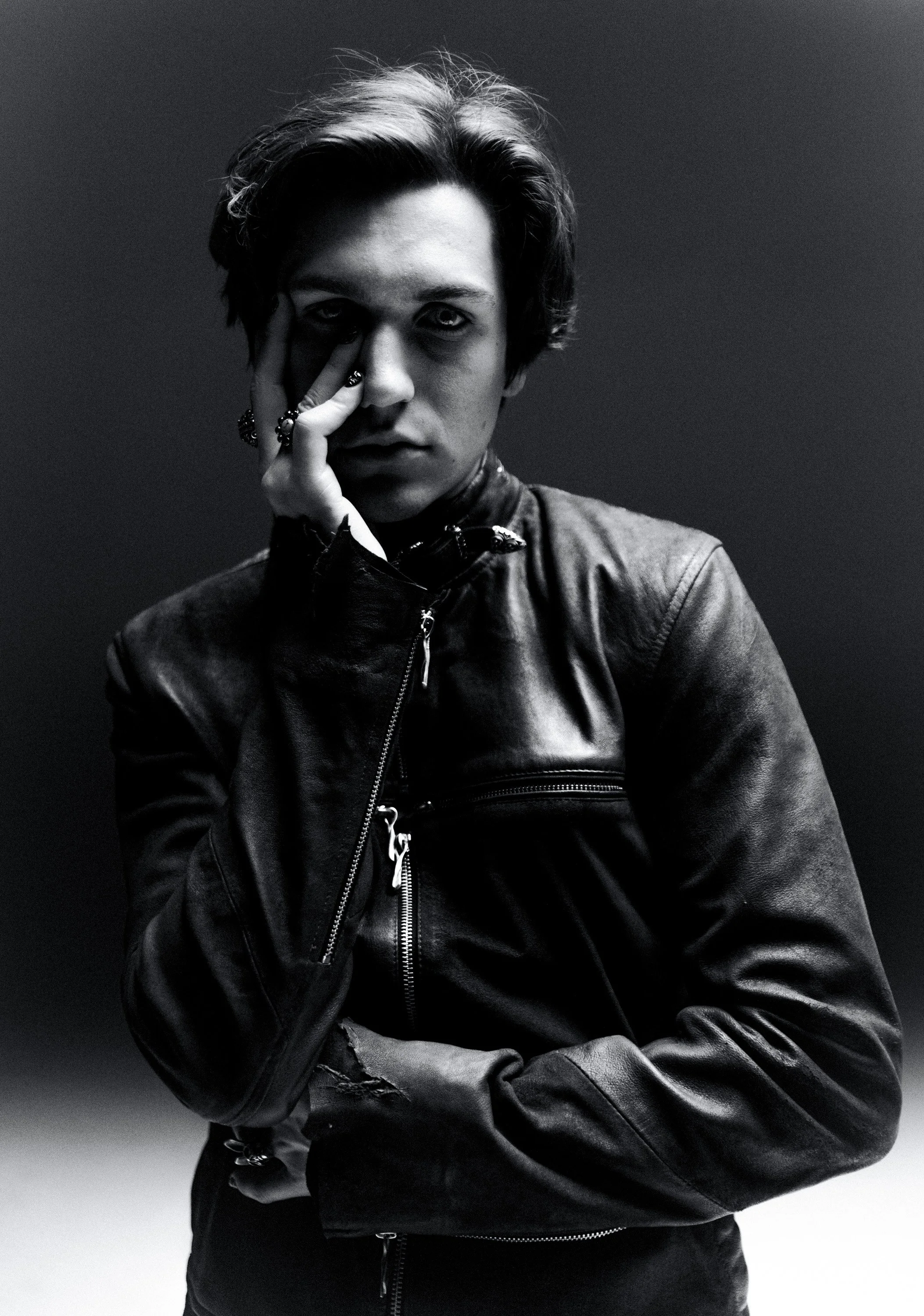 A black and white portrait of a young man with medium-length hair, wearing a leather jacket, with his left hand partially covering his face. He has jewelry on his fingers and an intense, serious expression.