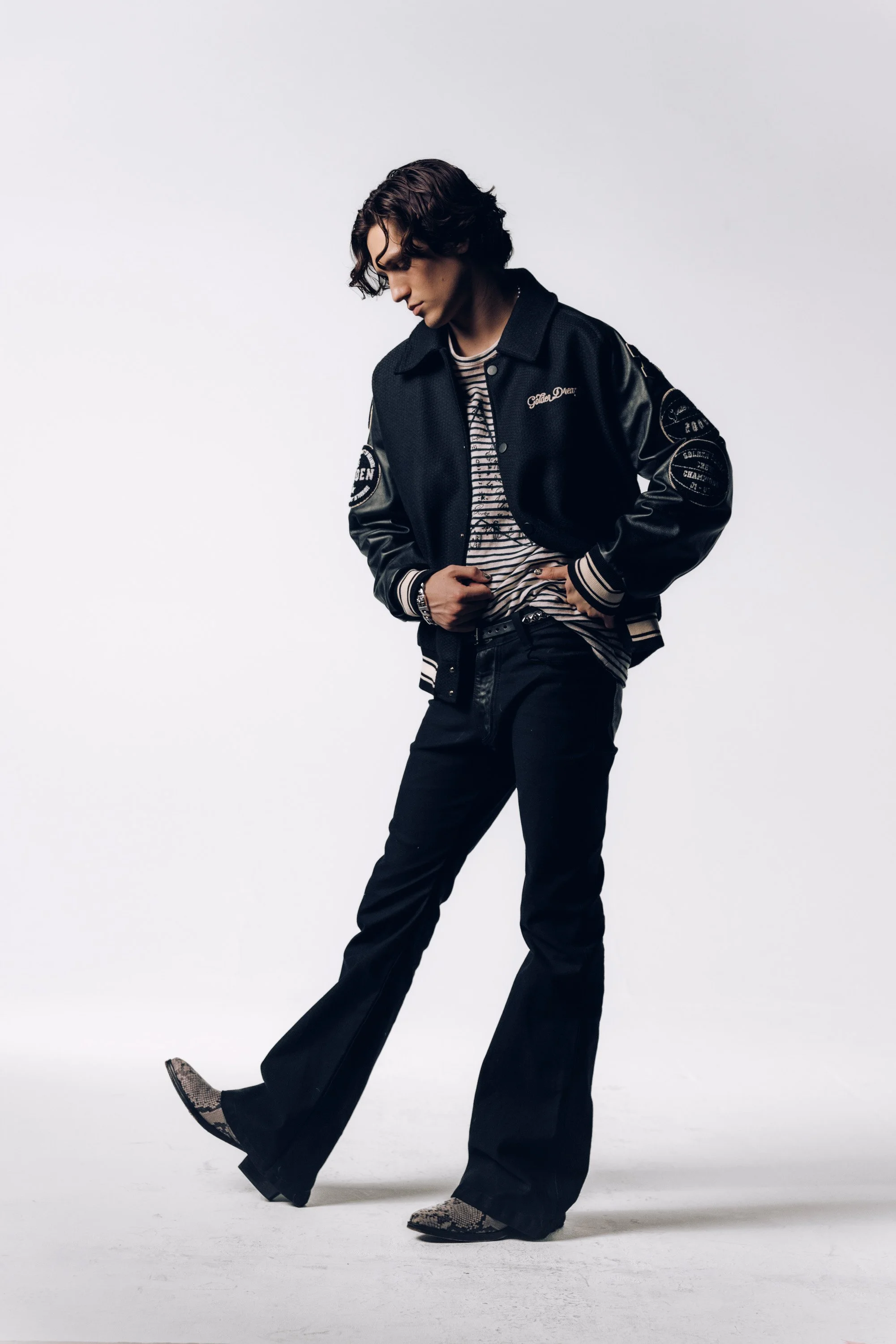 A young man with wavy, dark brown hair poses against a plain white background. He wears a black varsity jacket with patches, a striped shirt, black pants, and snake-patterned shoes. His gaze is downward, and one hand rests on his hip.