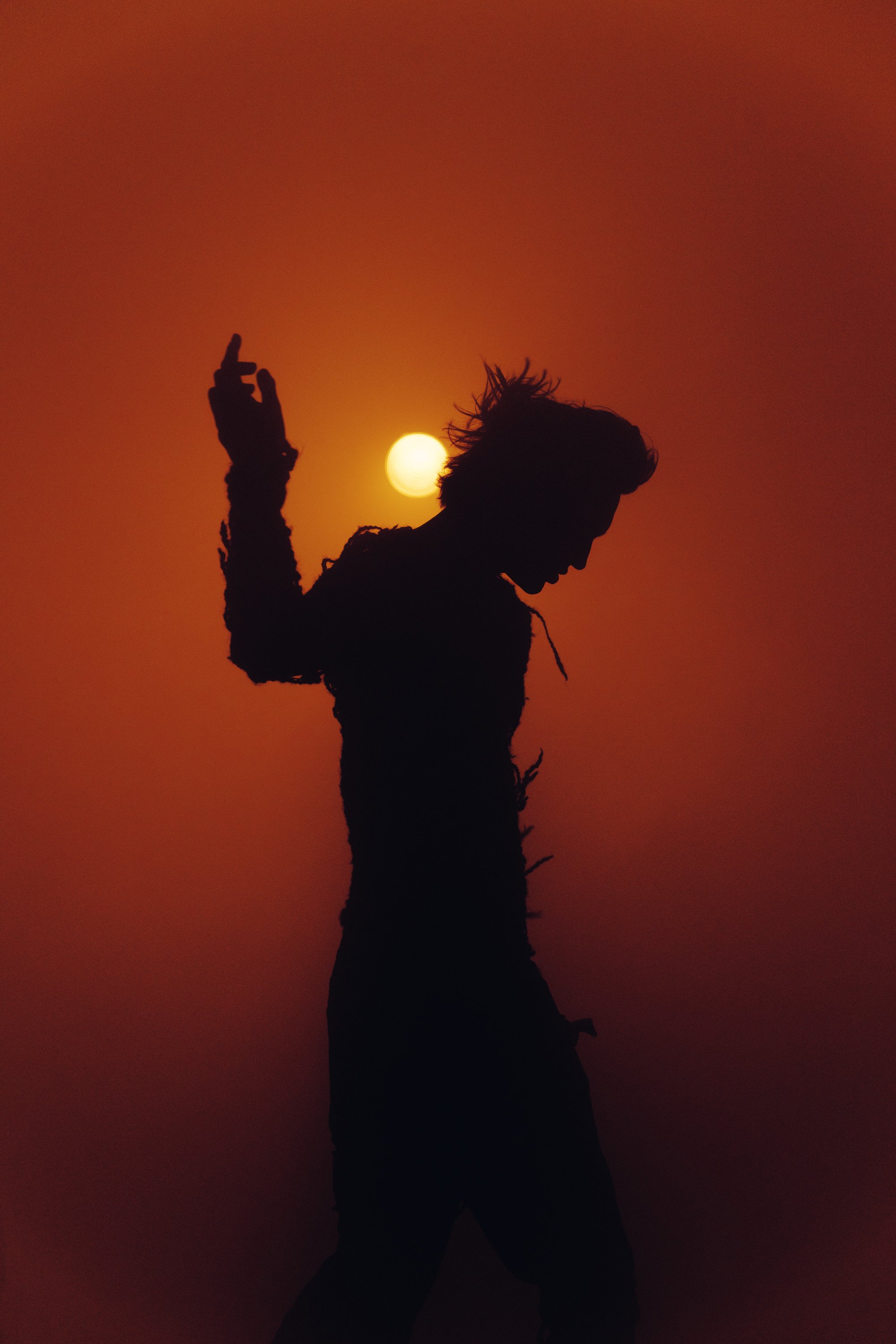 Silhouette of a person with spiky hair standing against an orange sunset background, with a bright sun near their head and one hand raised.