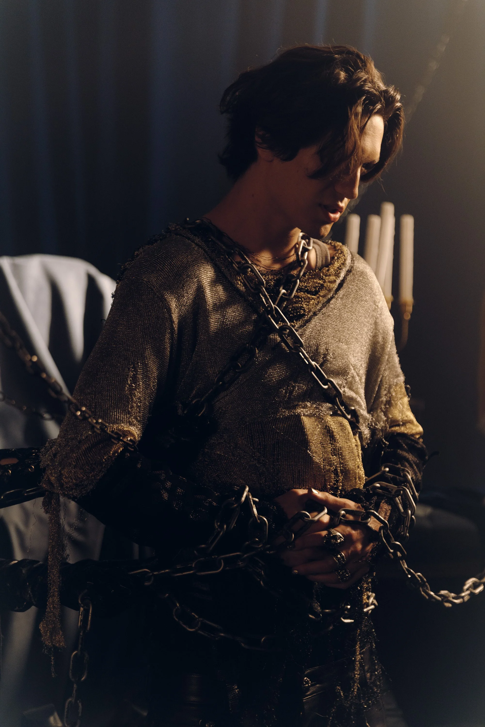 A young man with shoulder-length brown hair, wearing a shimmering gold top and chains, is chained up with large black metal links in a dimly lit room with a black curtain and candles in the background.