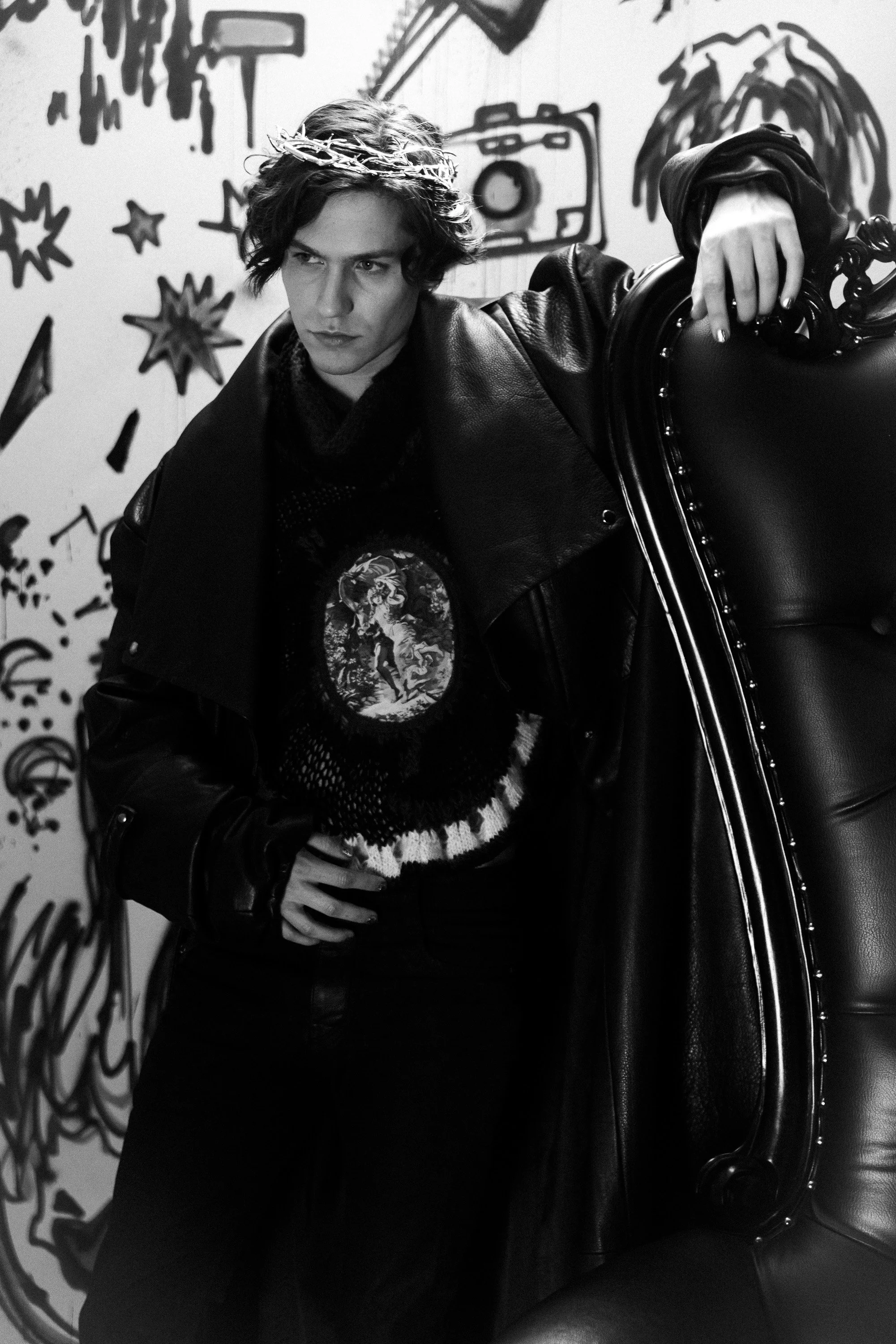 Black and white photo of a young man sitting in a vintage-style armchair. He has wavy hair, is wearing a leather jacket, a graphic T-shirt, and a crown-like headpiece. The background features a wall with abstract, graffiti-style drawings and doodles.