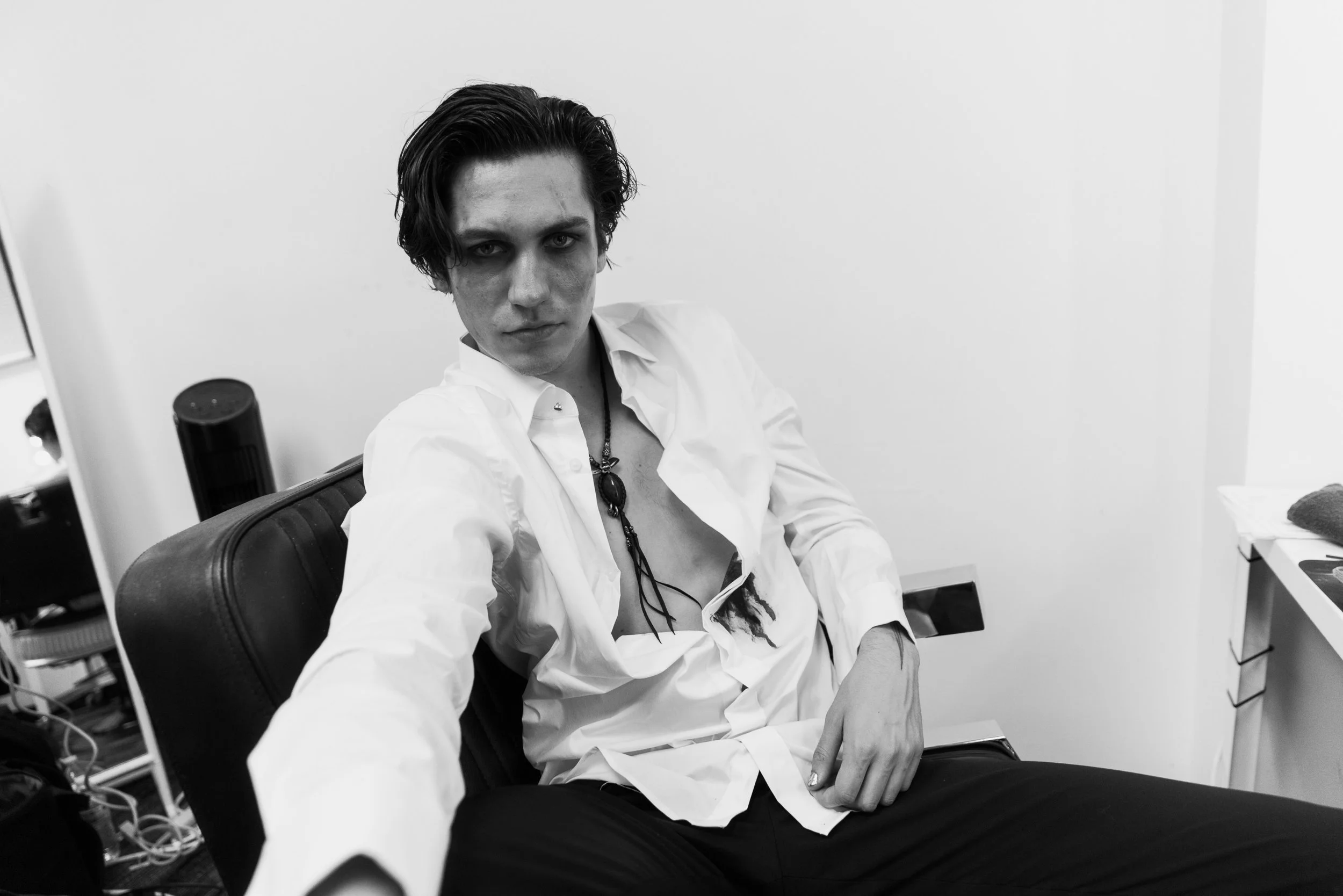 A black and white photo of a young man with dark, wavy hair sitting on a chair in a room with white walls. He is wearing a white shirt unbuttoned at the top, revealing a necklace, and has a serious expression on his face.