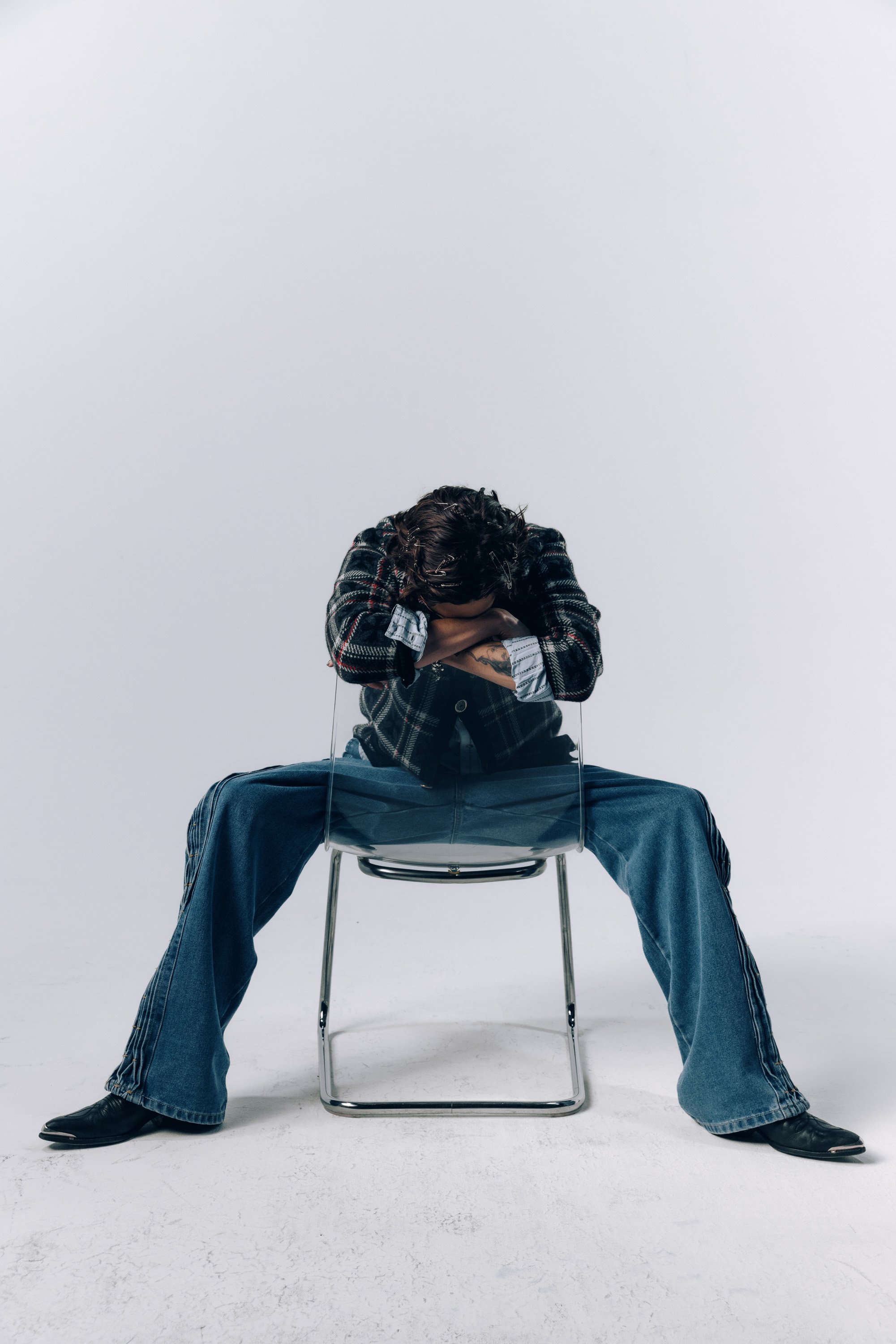Person sitting on a chair with legs wide apart, head resting on crossed arms on their knees, wearing a plaid jacket and jeans, in a plain white studio background.