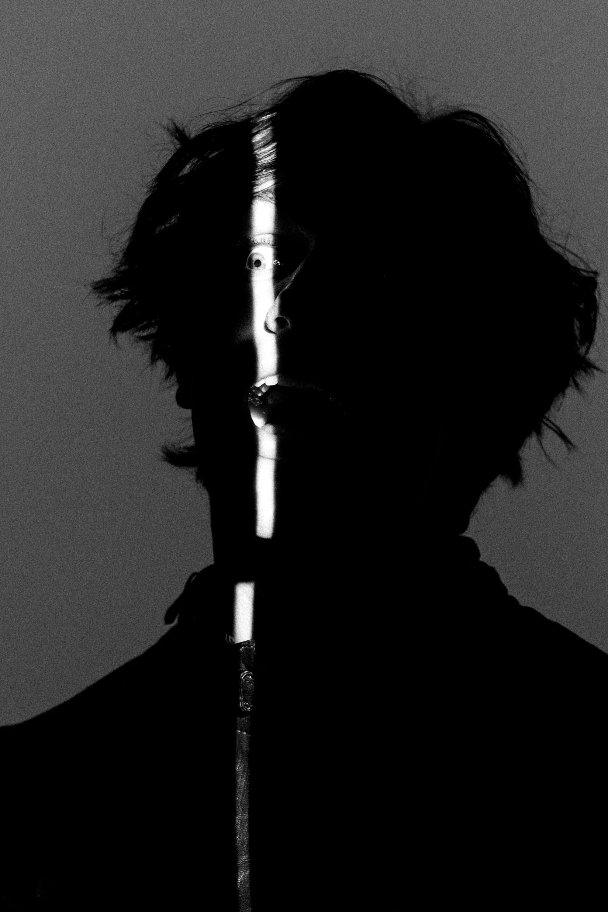 Black and white image of a person with messy hair, with half their face illuminated by a vertical light strip that runs down the middle of their face and body.